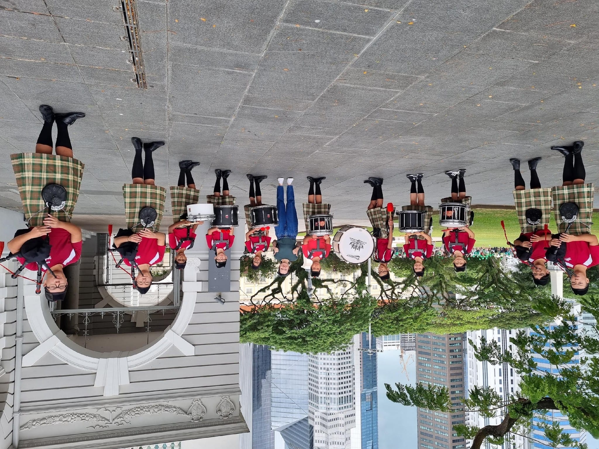 Upside-down photo of a marching band in red shirts and plaid kilts holding drums outdoors near buildings.