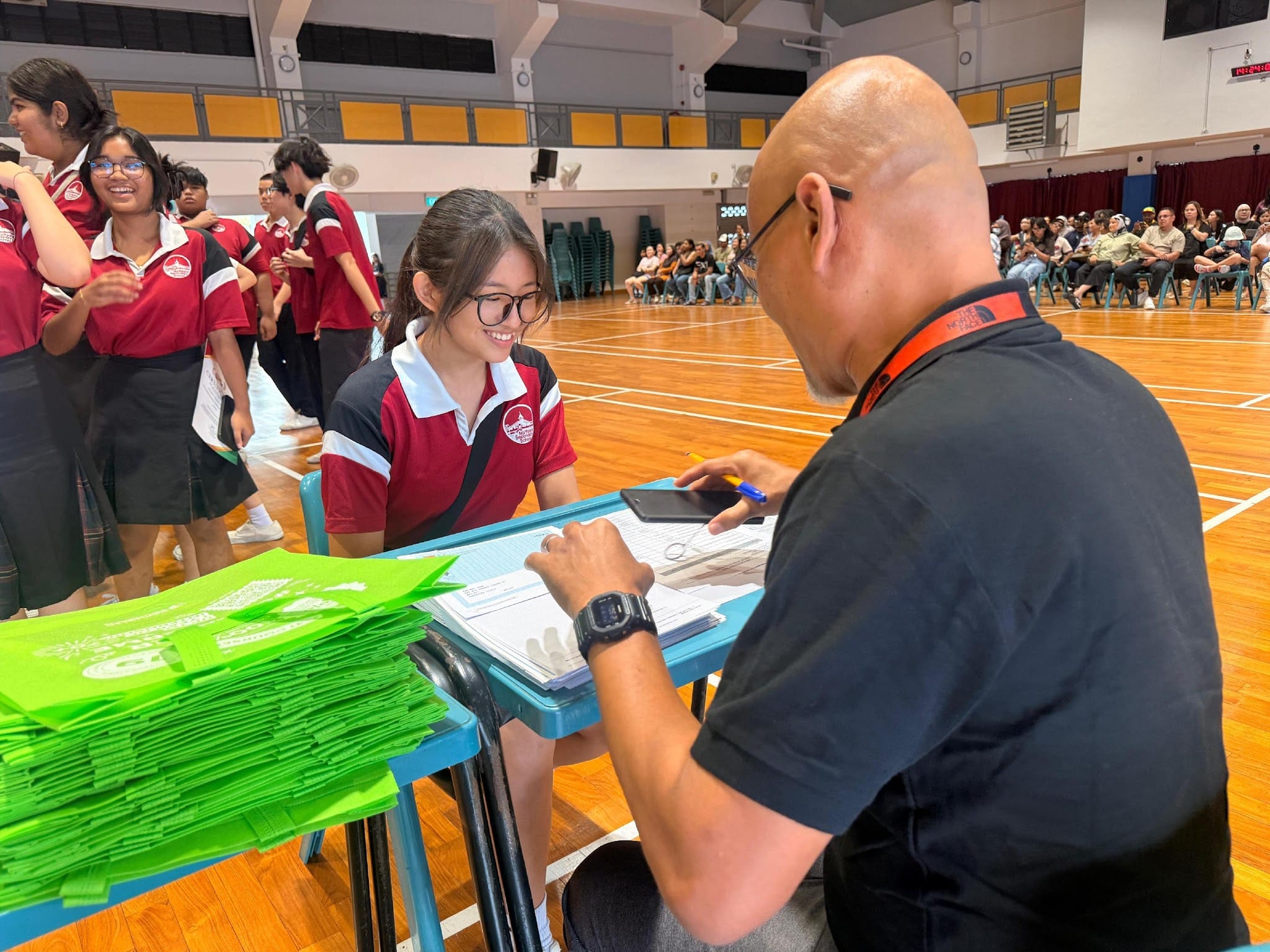 A smiling student in a red uniform assists a bald man signing documents at a table stacked with green bags in a school hall.