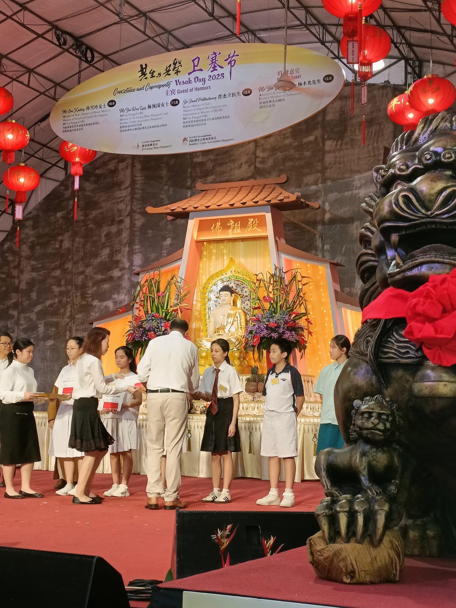 Children and adults stand on a stage at Vesak Day 2025 event, with a golden Buddha statue and red lanterns behind them.
