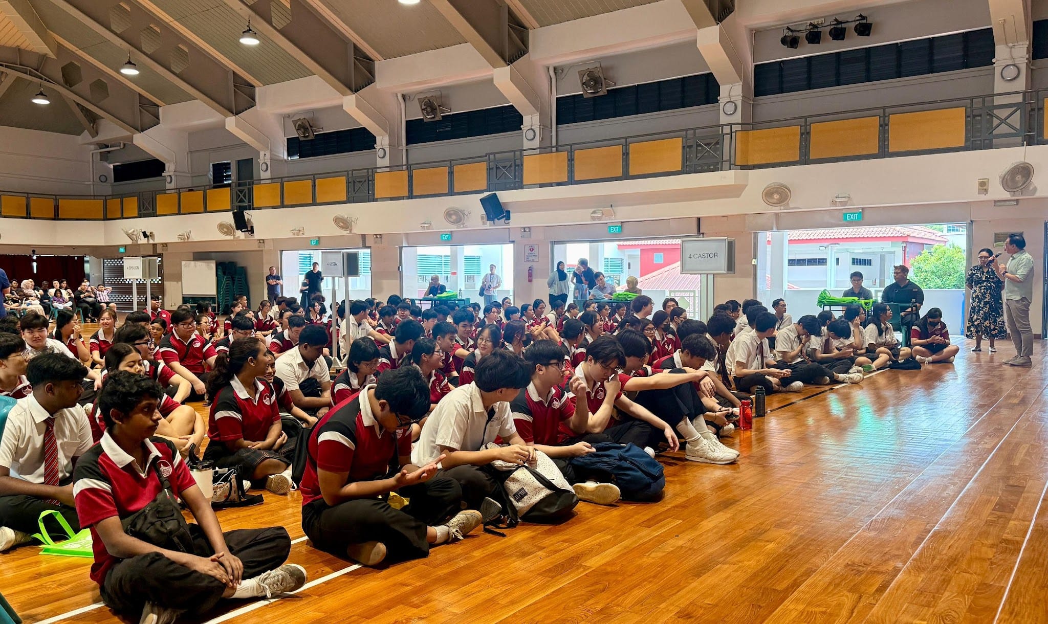 Students in red and white uniforms sit on a gymnasium floor facing speakers at the front of the hall.