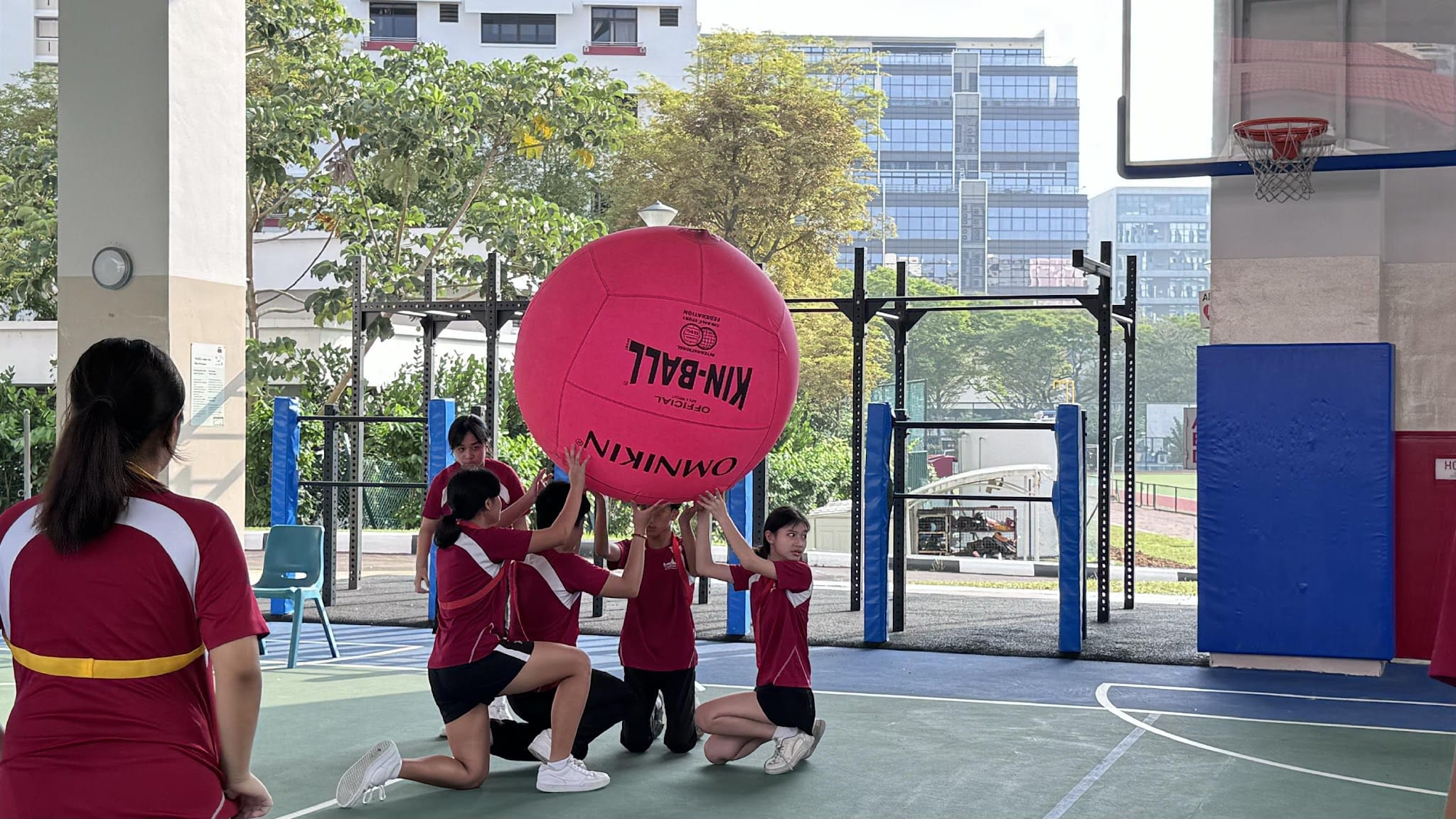 Students in red uniforms hold up a large pink Kin-Ball on an outdoor court with buildings in the background.