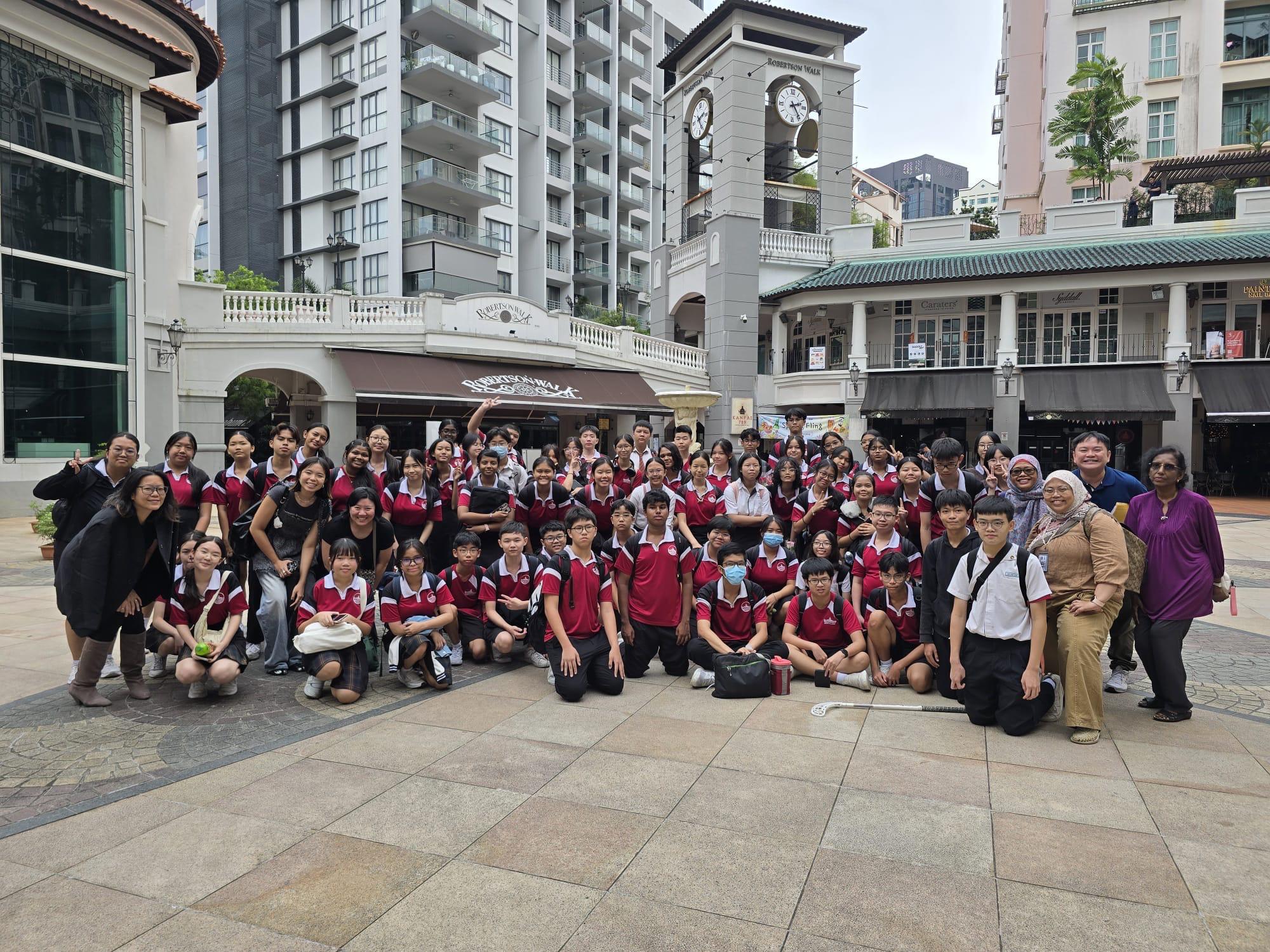 Large group of students in red and white uniforms posing outdoors at Robertson Walk with a clock tower behind them.