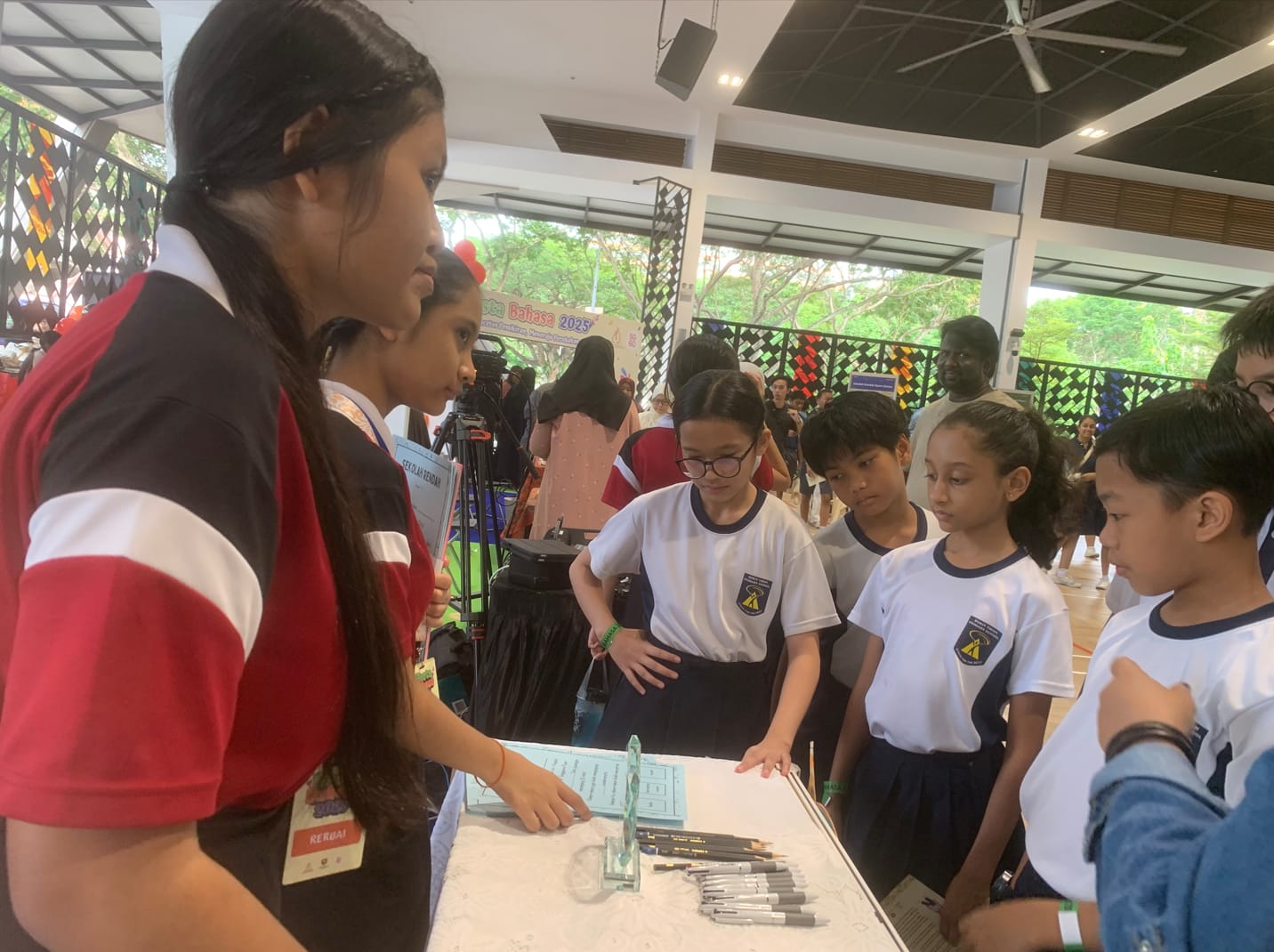 Students in uniforms gather around a table with pens and papers at an indoor school event or fair.