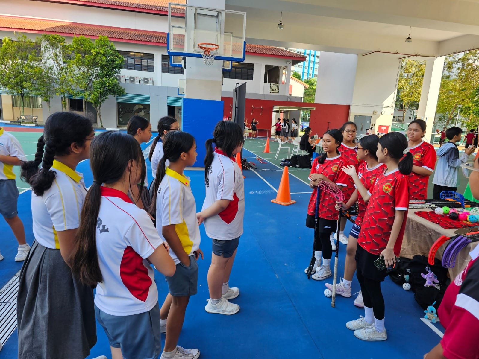 Students in white and red jerseys gather on a blue outdoor basketball court near sports equipment and orange cones.