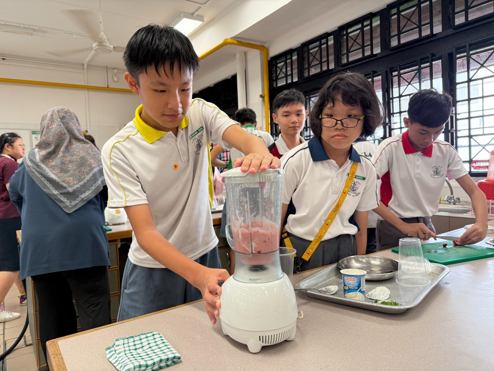 Students in school uniforms use a blender with pink contents during a cooking class activity.