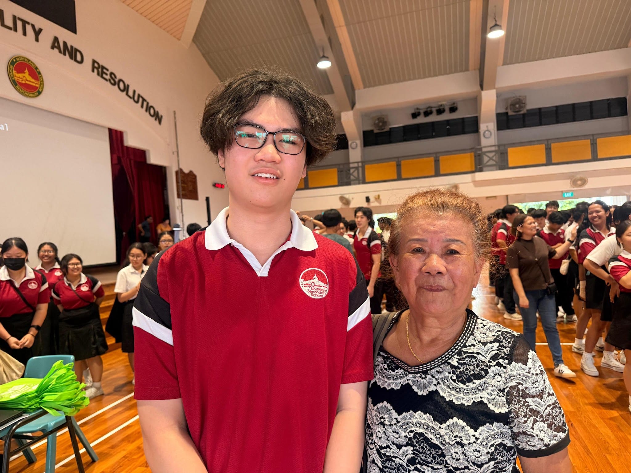 A teenage boy in a red school uniform with glasses stands beside an elderly woman in a school hall.