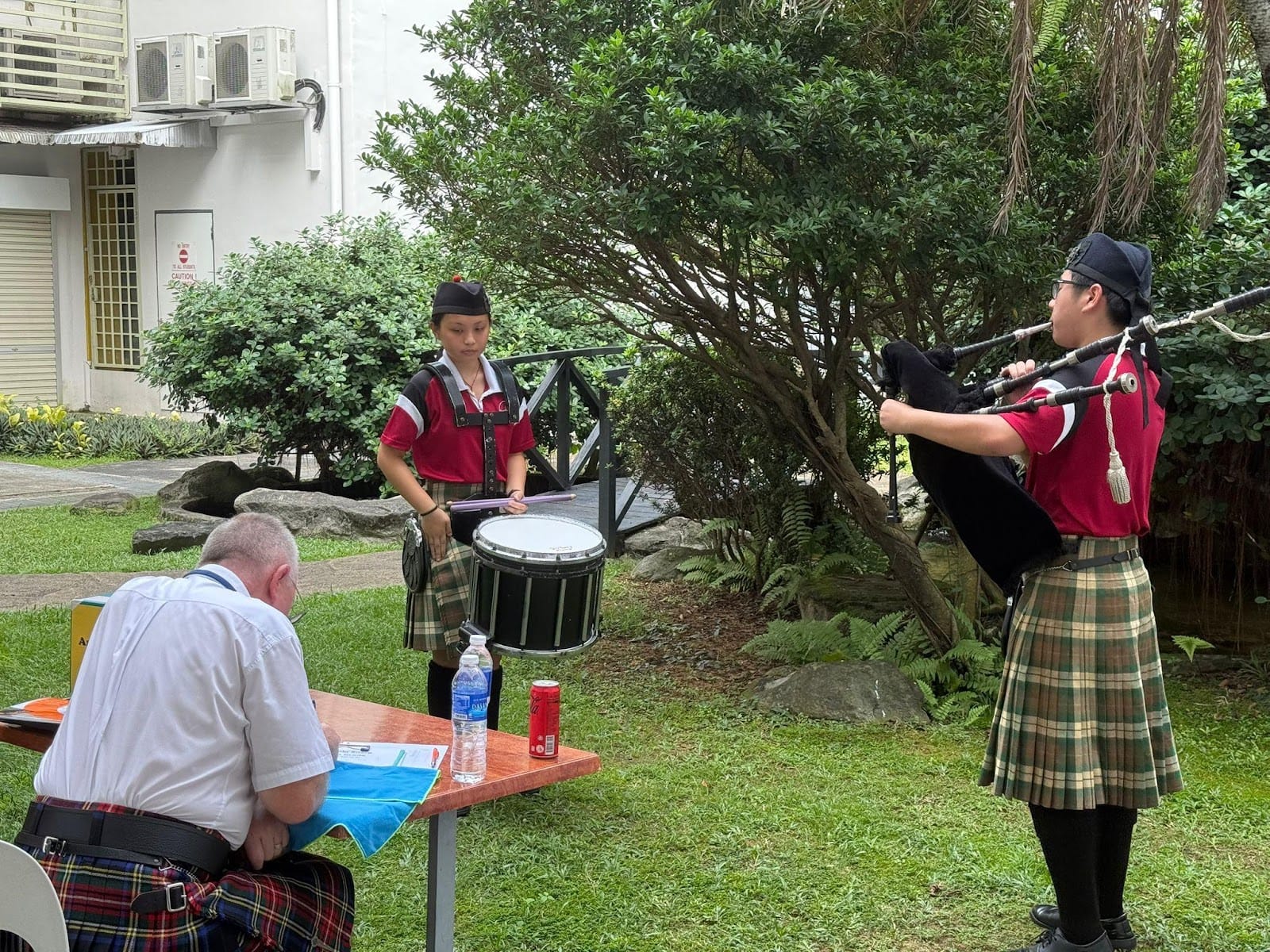 Two young people in kilts and berets play bagpipes and snare drum outdoors while a man sits at a table.