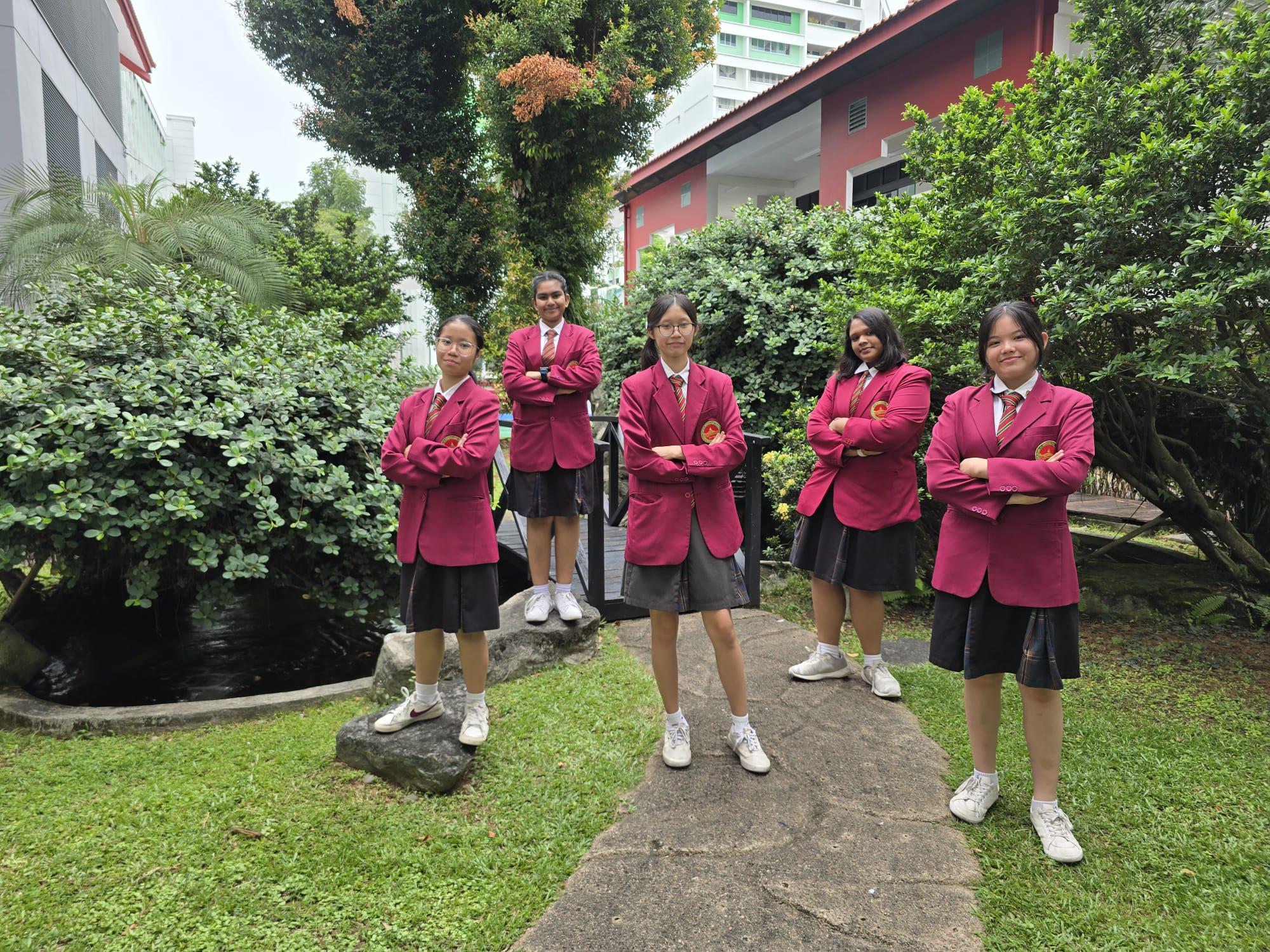 Five girls in matching red blazers and gray skirts stand with arms crossed in a green school garden.