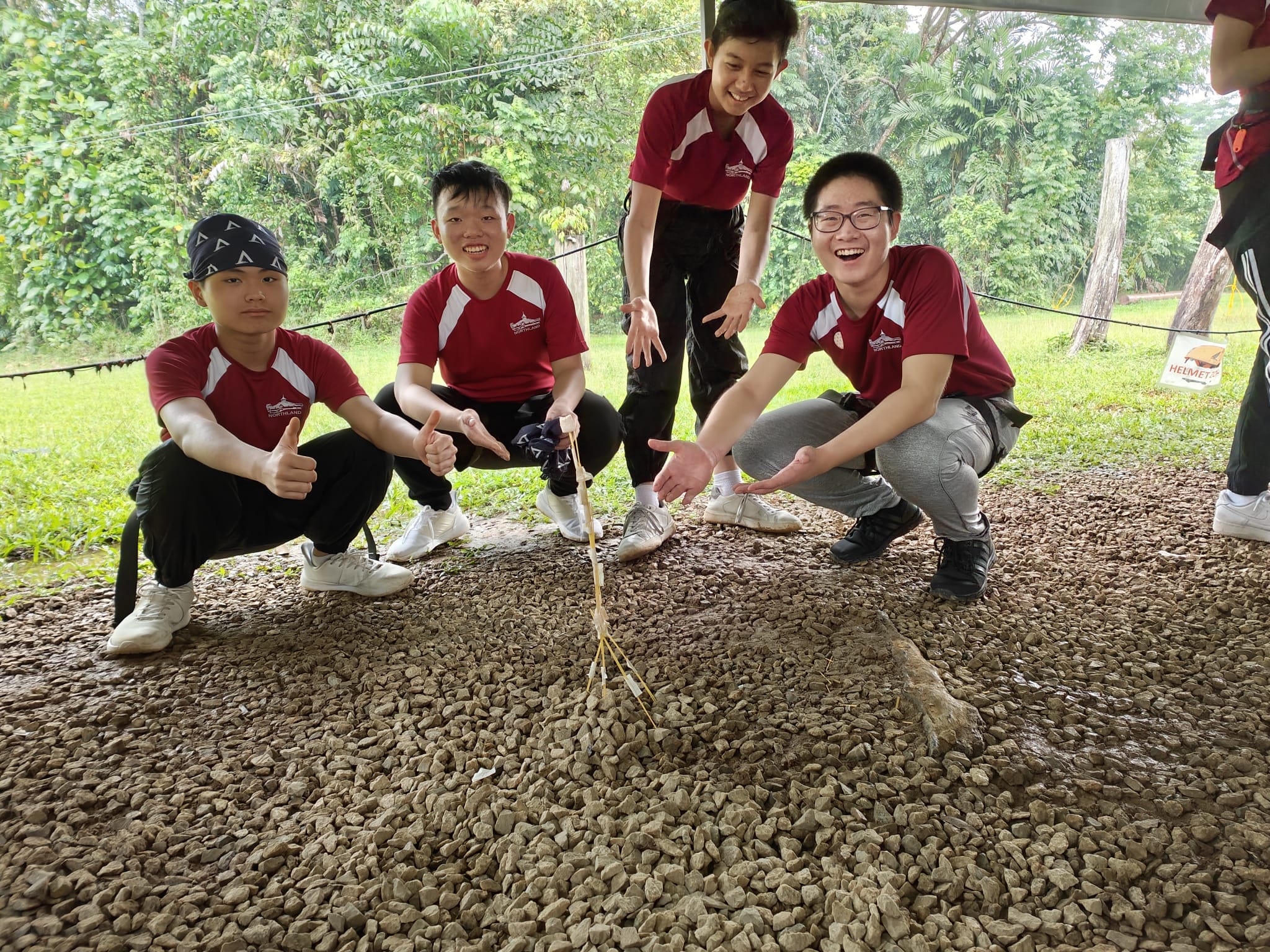 Four youths in red uniforms crouch on gravel, smiling and pointing at a small stick structure they built outdoors.