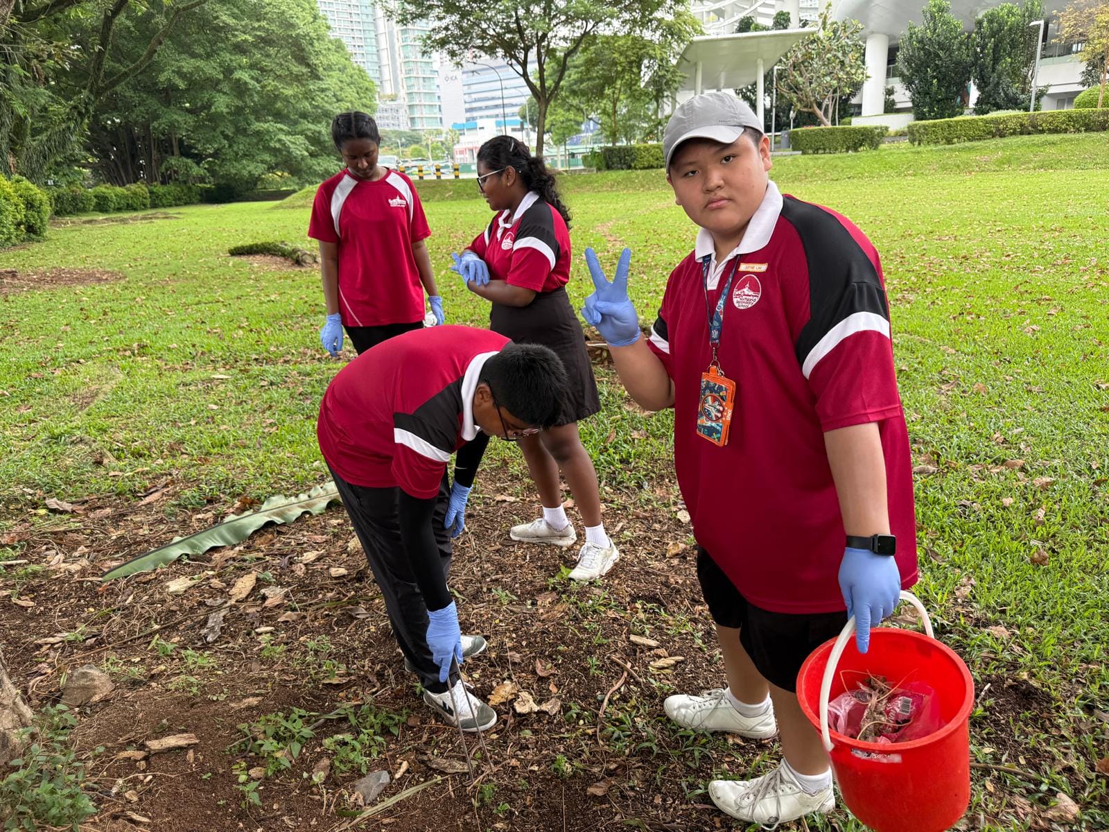 Four students in red uniforms and blue gloves clean up a park, one holding a red bucket and making a peace sign.