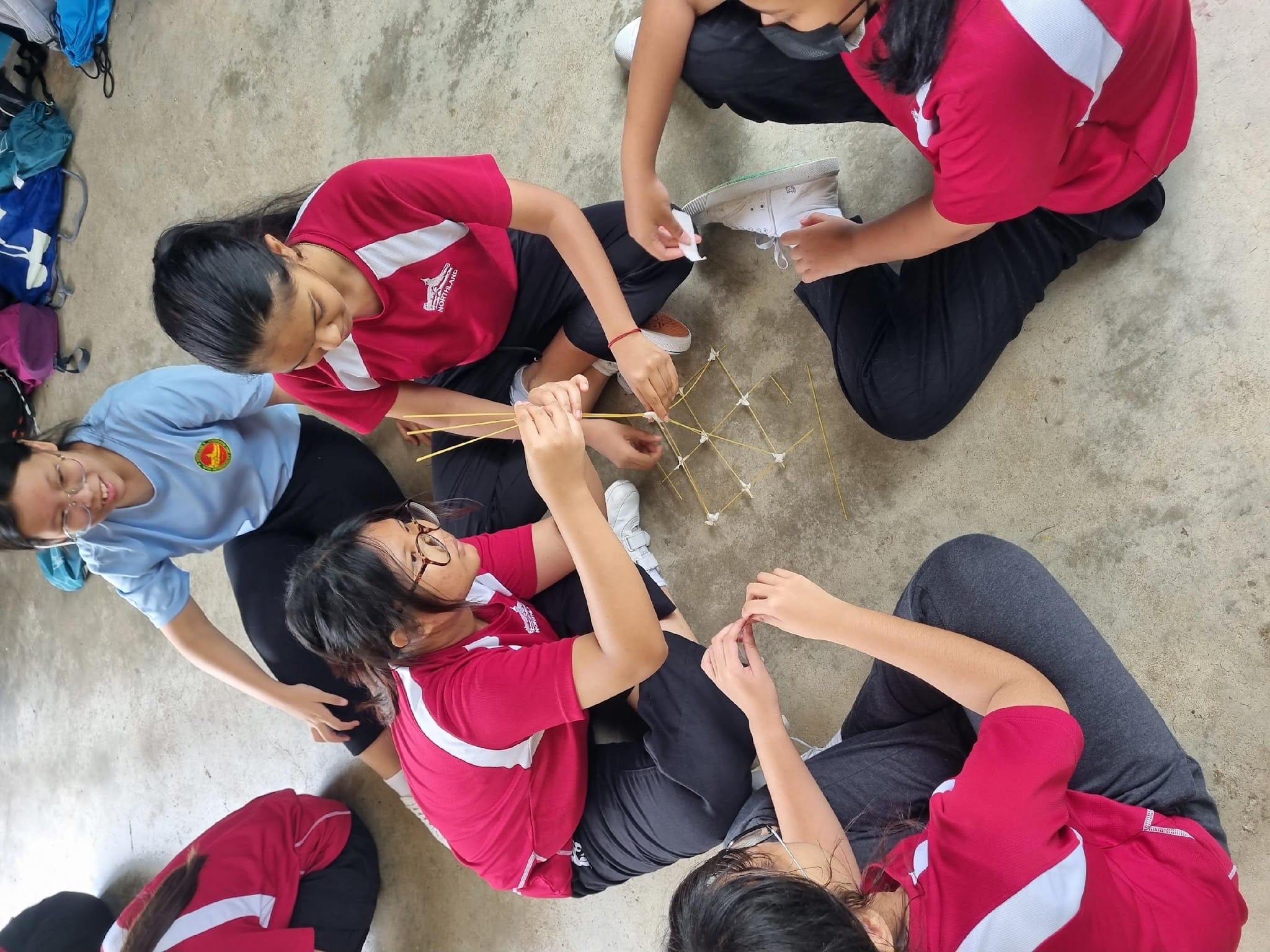 Students in red uniforms sit on the ground building a structure using sticks and marshmallows together.