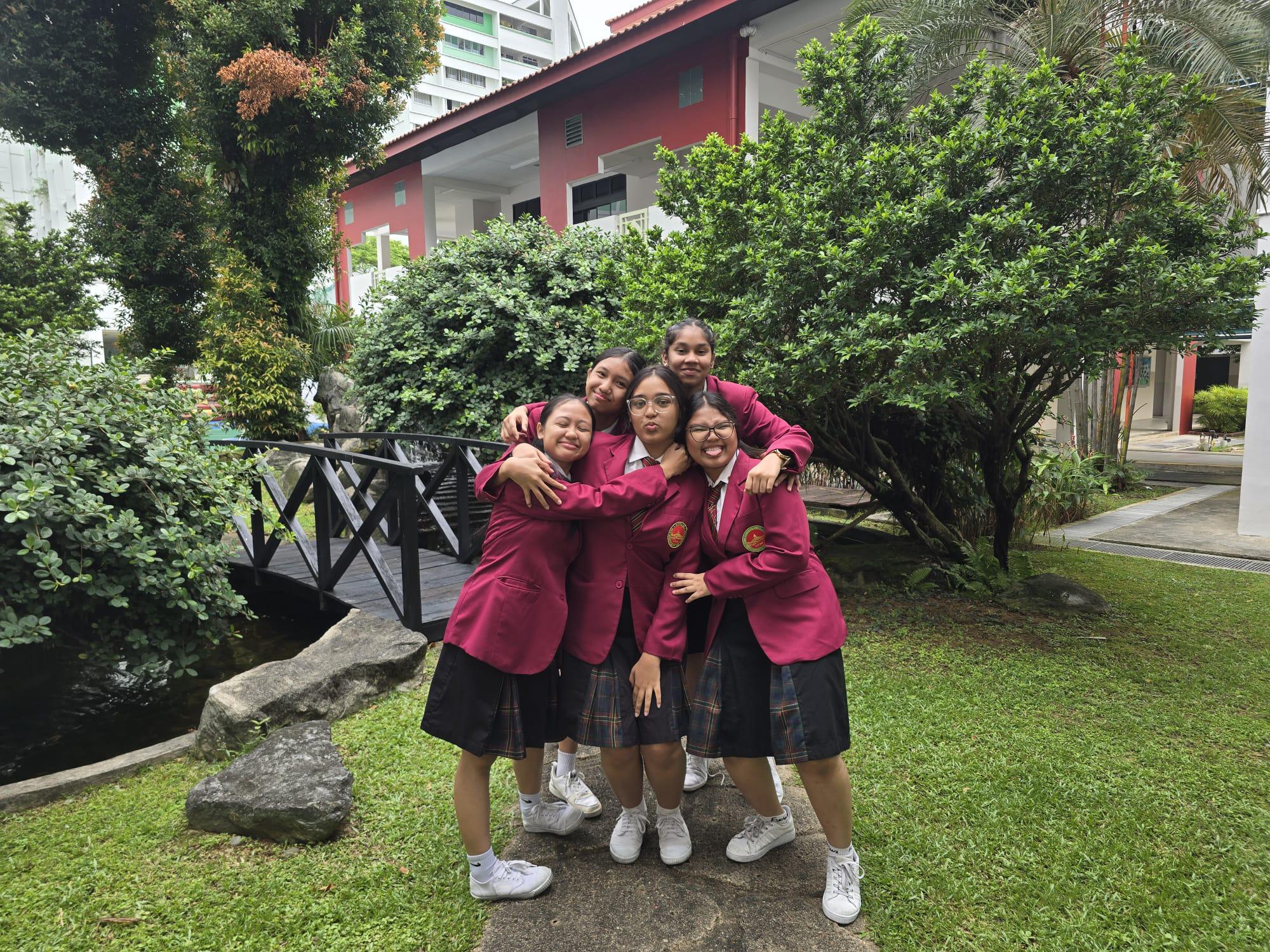 Five girls in matching maroon blazers and plaid skirts pose hugging outdoors near a small wooden bridge.