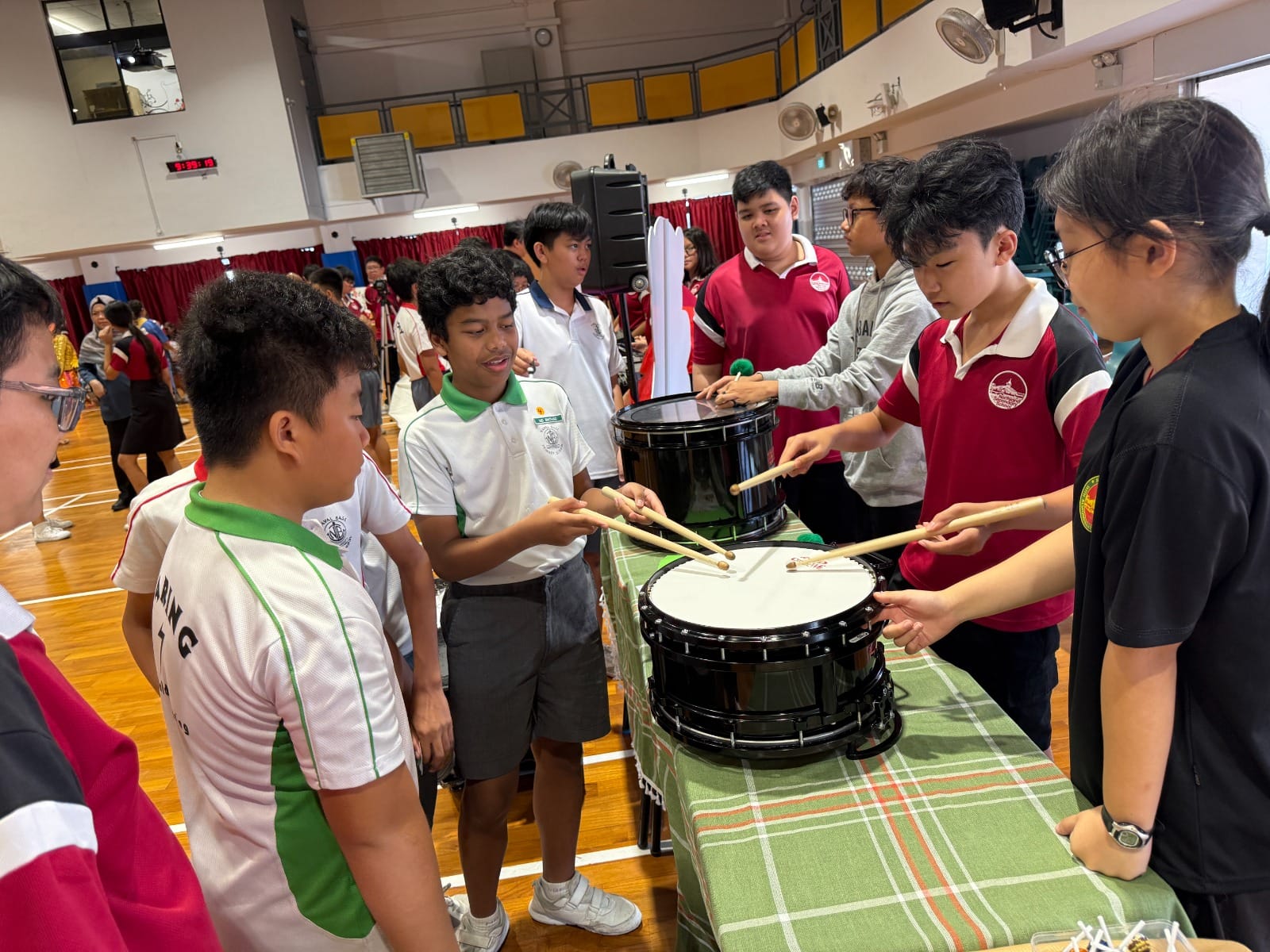 Students in school uniforms try playing snare drums with drumsticks at a table in a school hall.