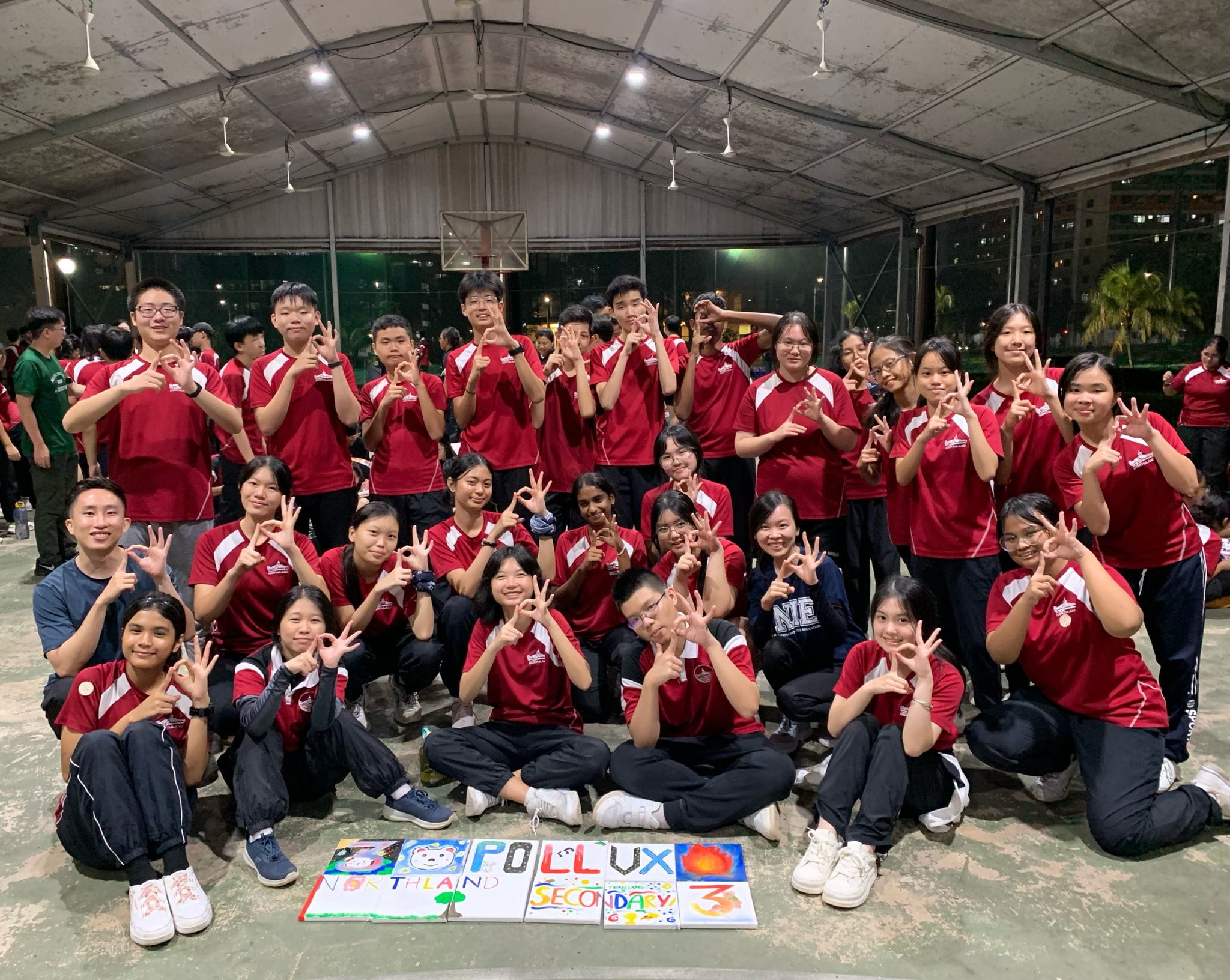 A large group of students in red uniforms pose with hand signs beside colorful signs reading Pollux Northland Secondary 3.