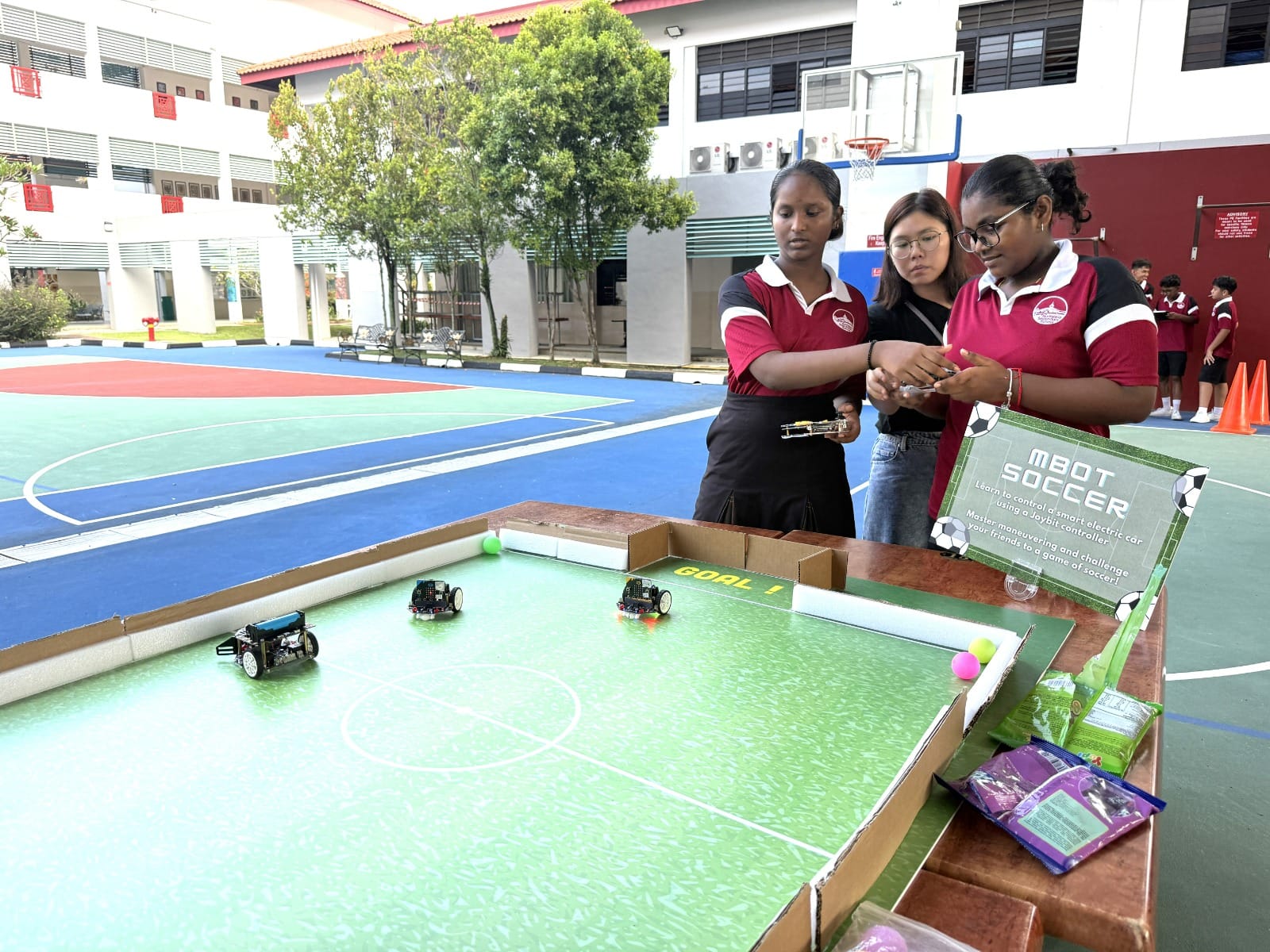 Three students stand around a cardboard mBot Soccer field with small robots, at a school outdoor court.