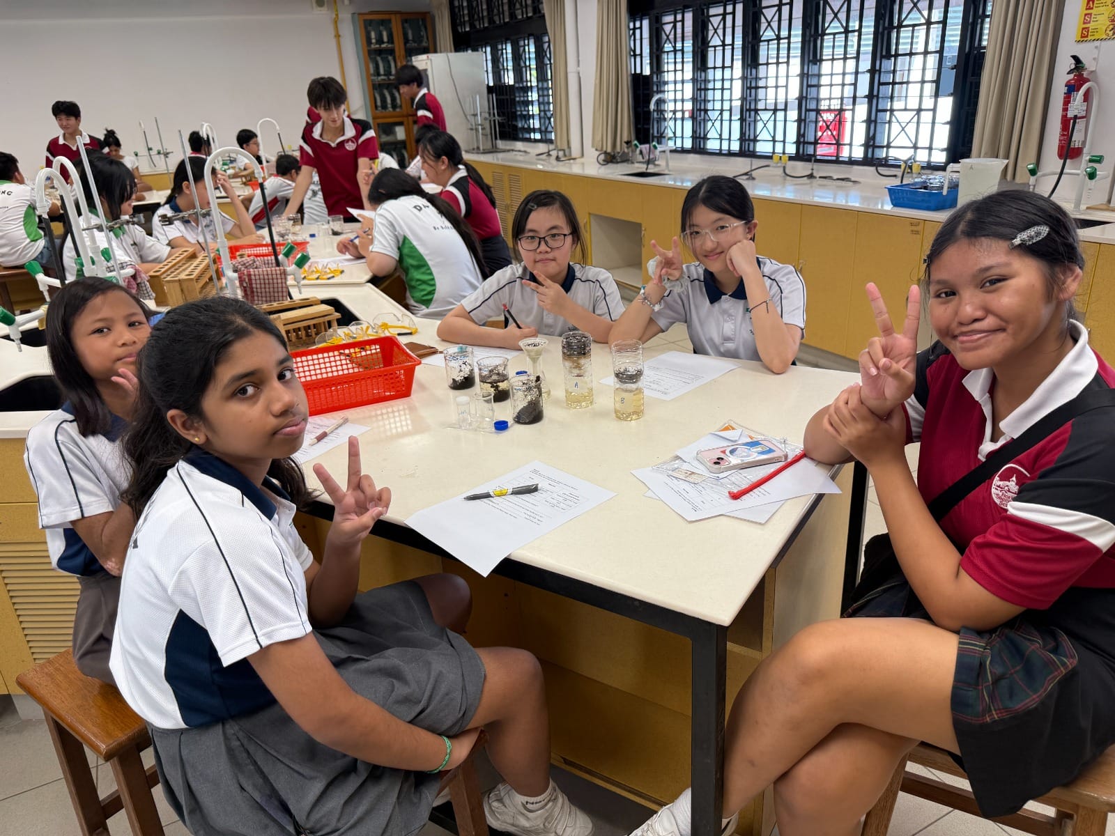 Four smiling schoolgirls in uniforms pose with peace signs at a lab table with beakers during a science class.