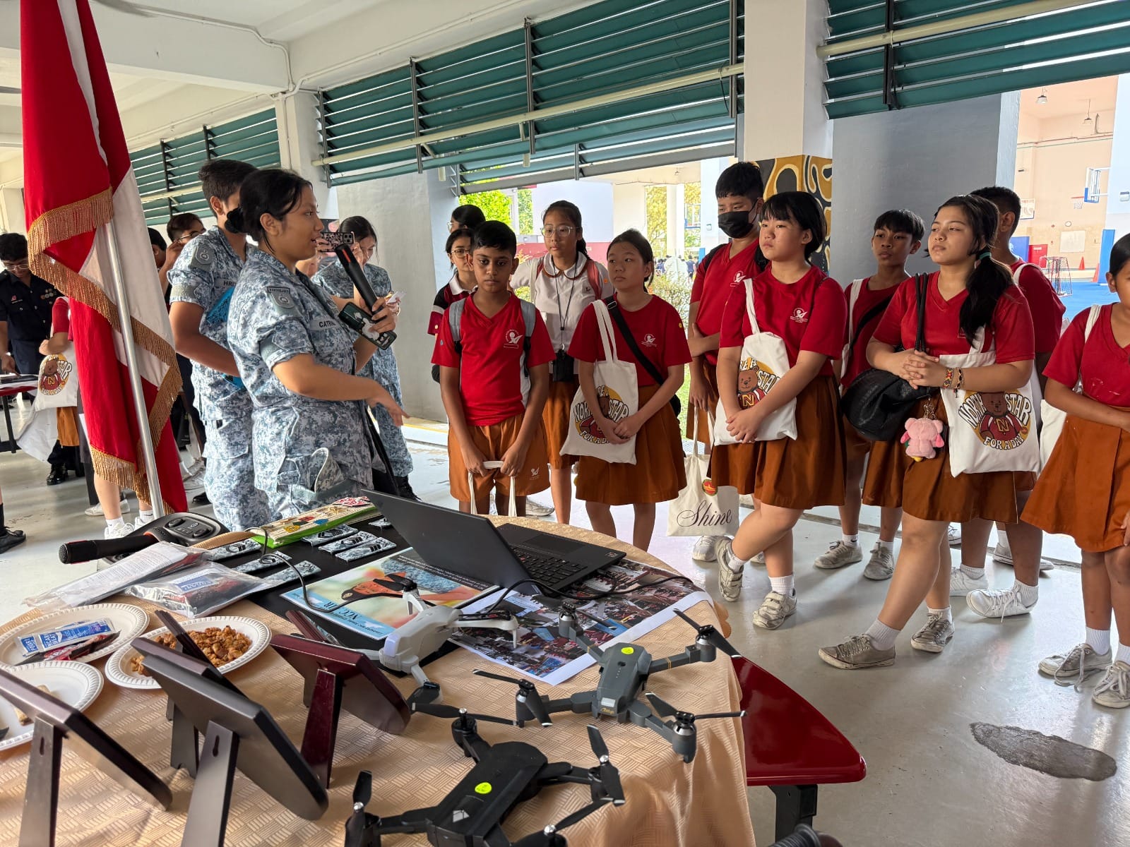 A uniformed officer presents drones and equipment on a table to a group of students in red uniforms at a school event.