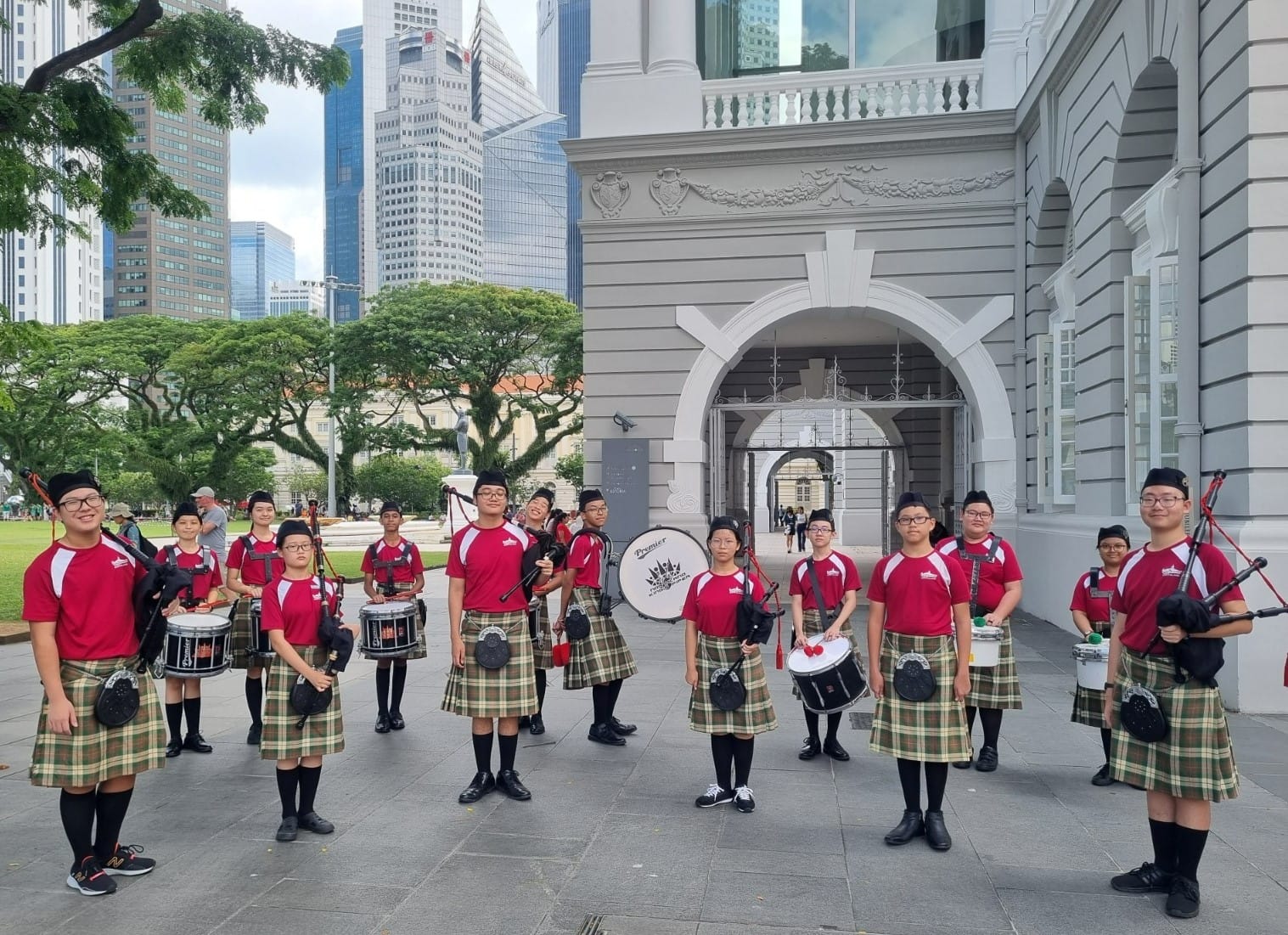 Youth pipe band members in red shirts and tartan kilts holding bagpipes and drums outside a colonial building.