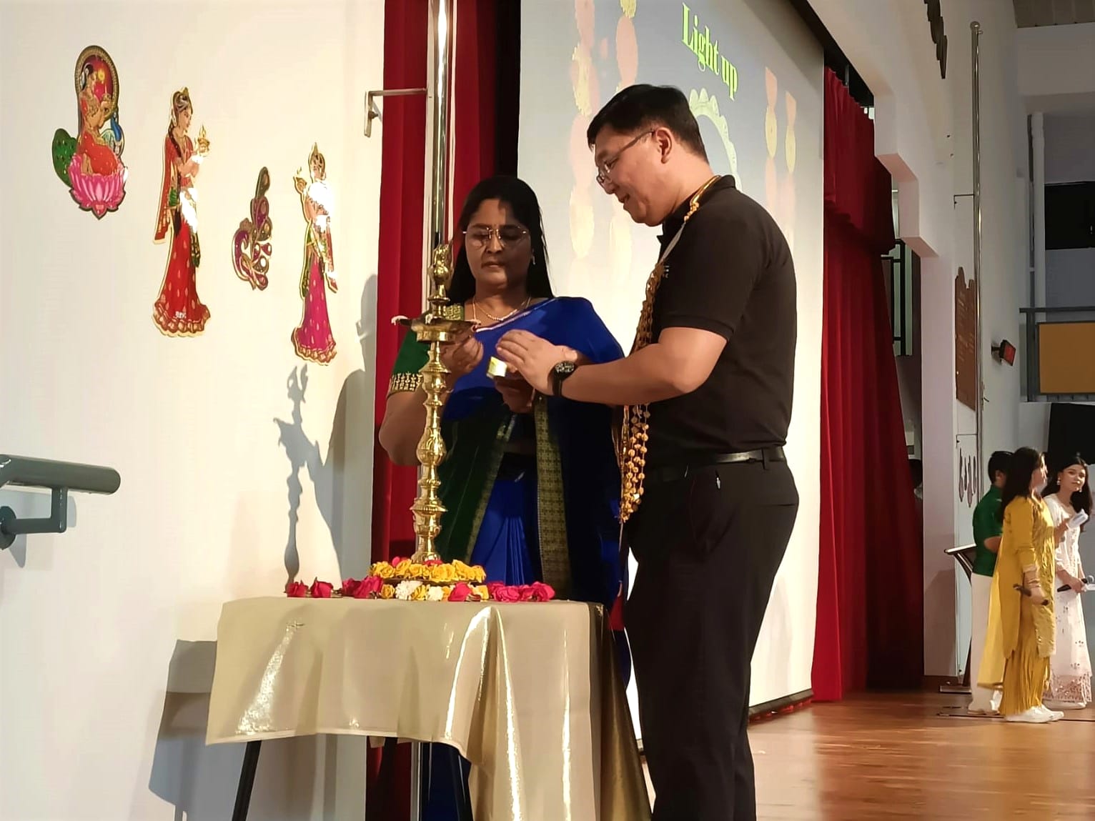 Two people light a traditional brass oil lamp on a flower-decorated table at a Diwali celebration on stage.