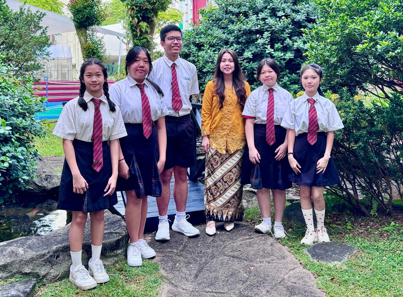 Six young people pose outdoors; five wear white shirts with red ties, and one wears a gold traditional outfit.