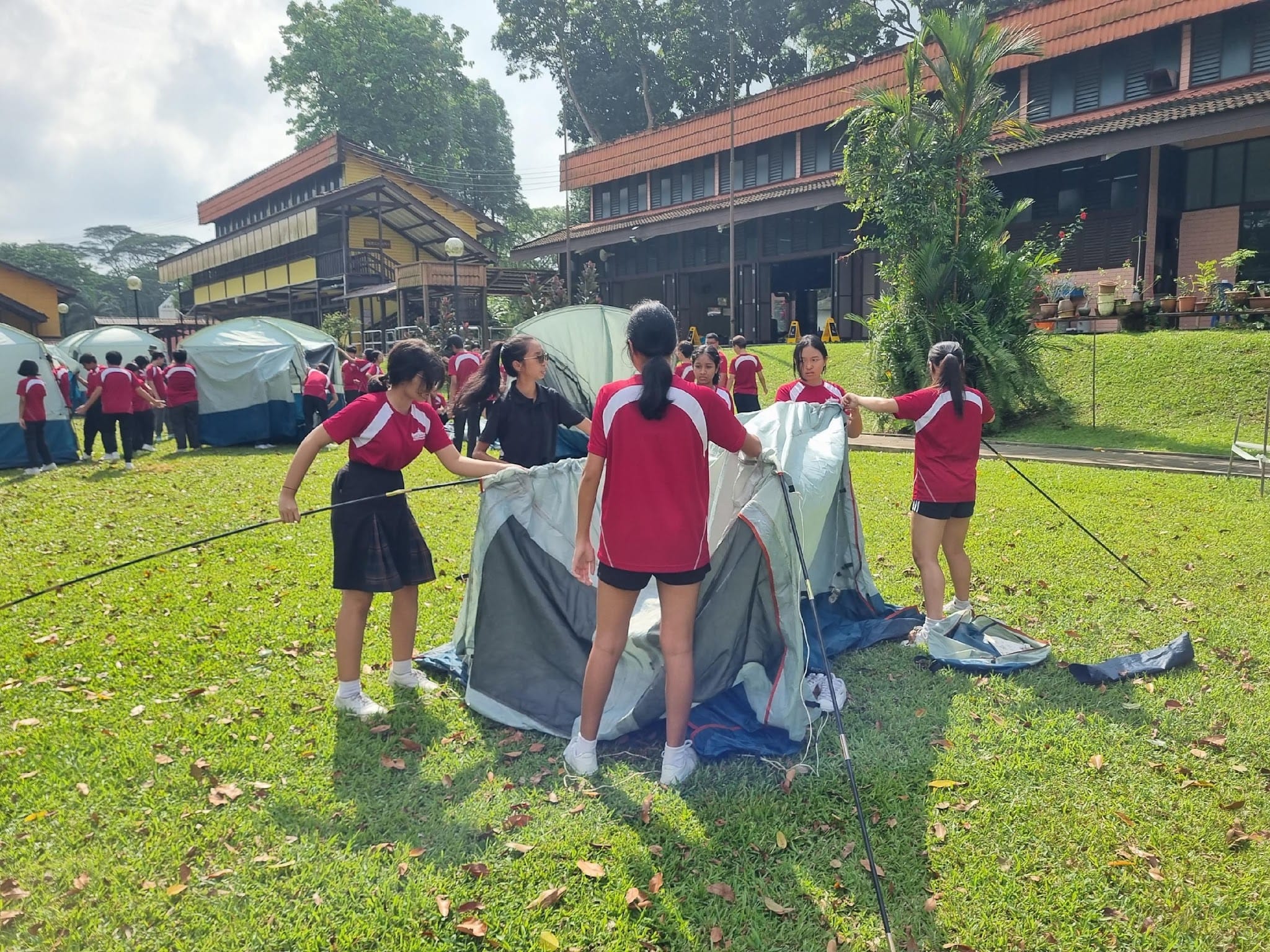 Students in red uniforms set up tents on a grassy field near buildings during an outdoor camp activity.