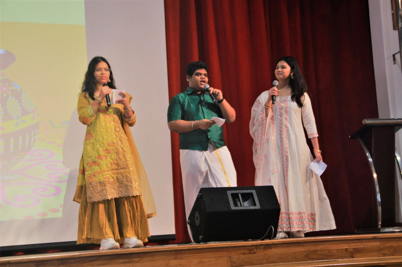 Three young people in traditional Indian attire hold microphones and papers on a stage with red curtains.