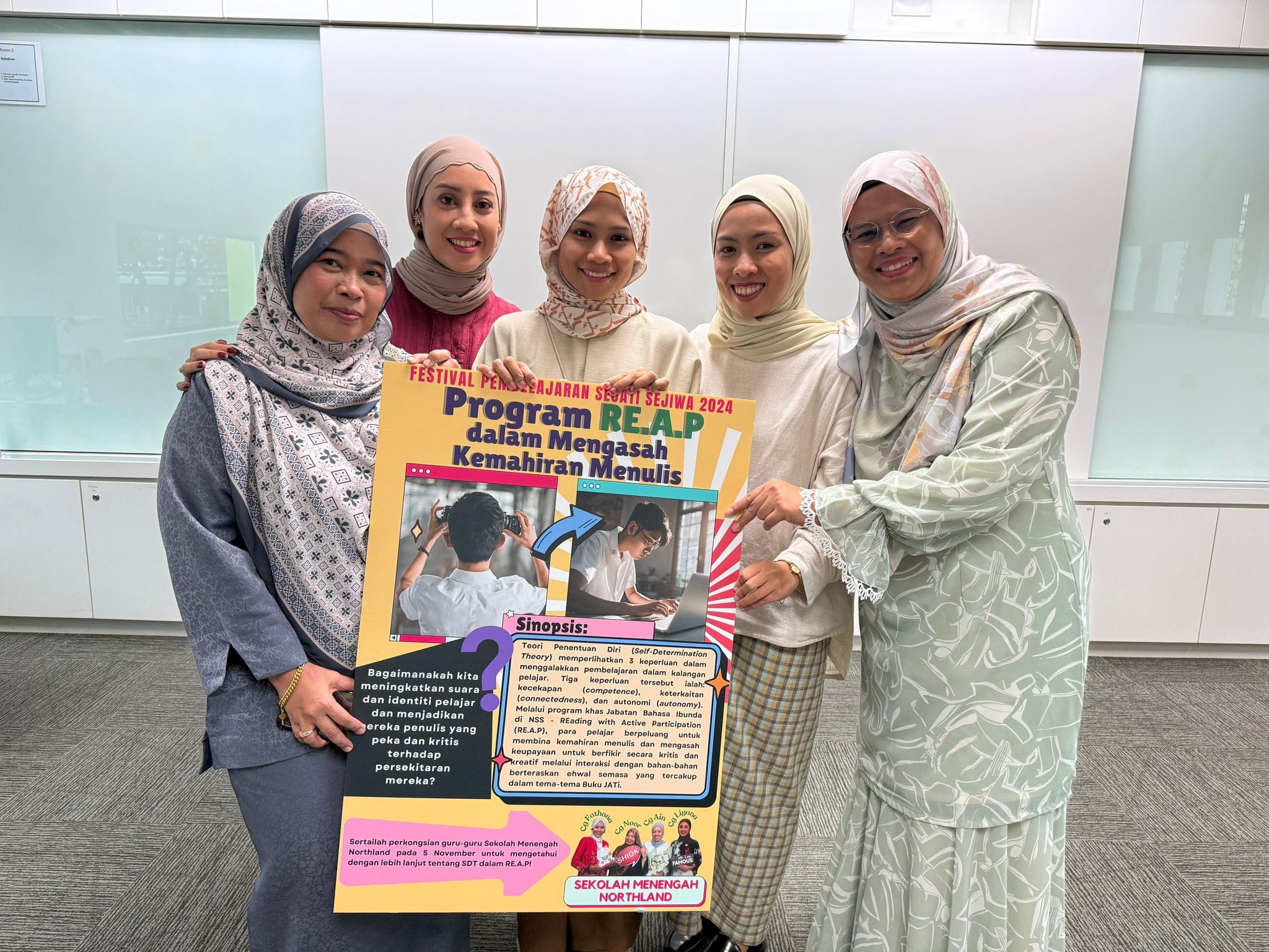 Five smiling women wearing hijabs hold a colorful Malay-language poster about Program RE.A.P writing skills.