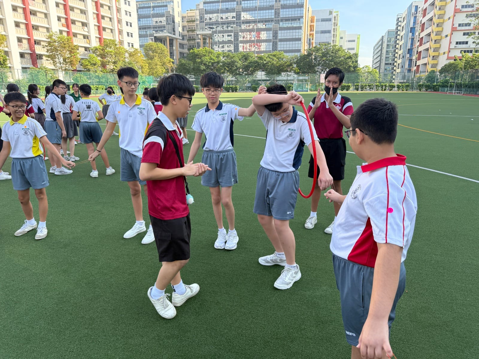 Students in school uniforms play a rope activity on a green outdoor field surrounded by tall buildings.
