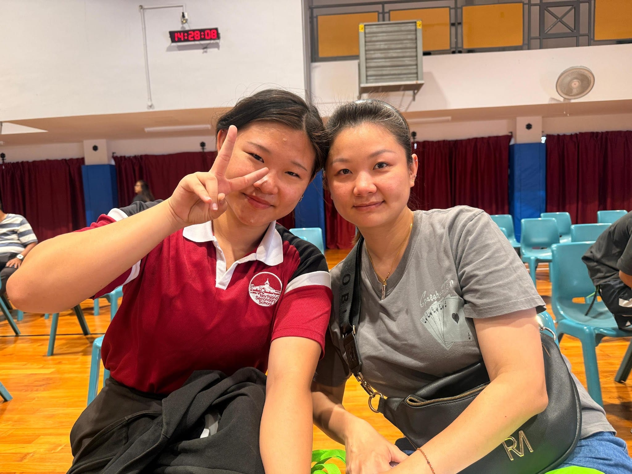 Two young women smile in a school hall, one in a red uniform making a peace sign, the other in a gray shirt.