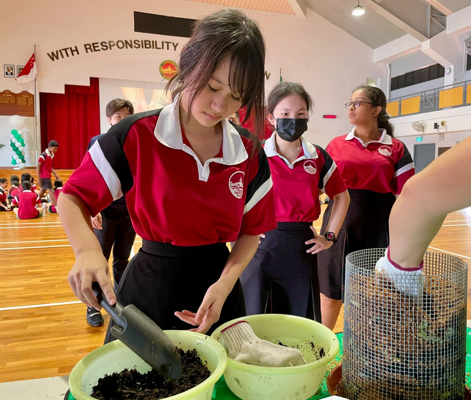 Students in red uniforms handle soil with a trowel and bowl in a school hall with a responsibility banner.