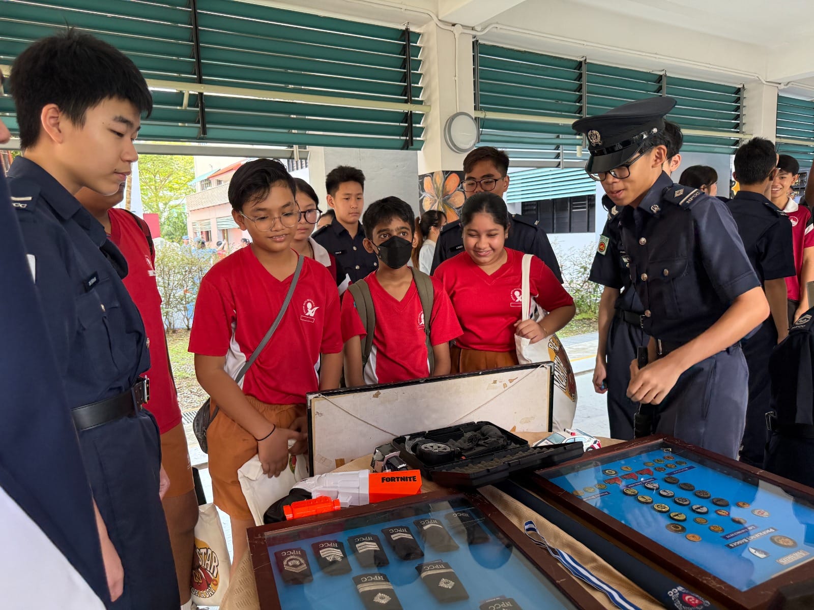 Students in red shirts gather around display cases of badges and equipment while a uniformed officer explains the items.