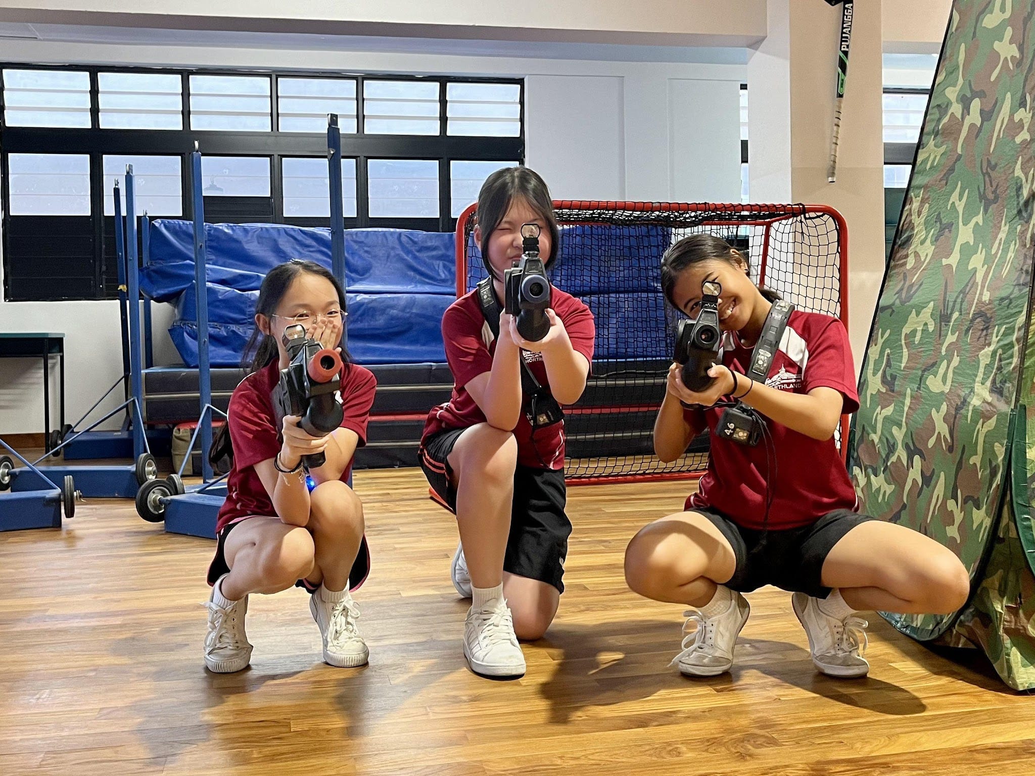 Three girls in red shirts crouch on a wooden floor, each aiming a laser tag gun toward the camera indoors.