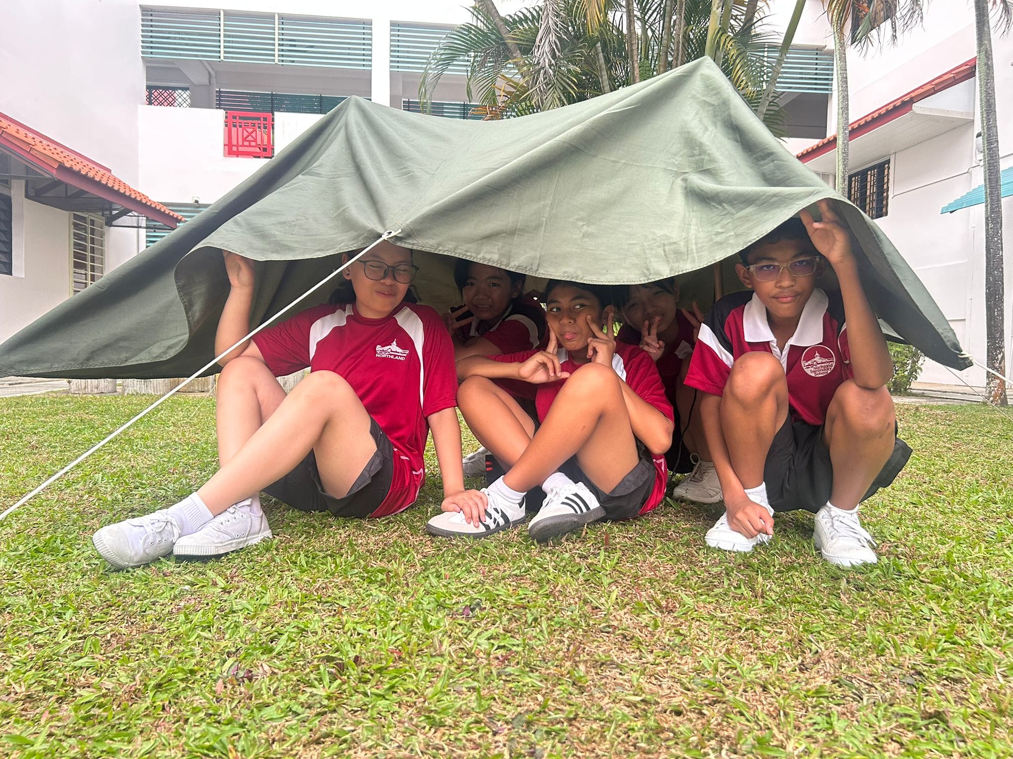 Five students in red school uniforms sit together under a green tarp shelter on a grass field, posing with peace signs.
