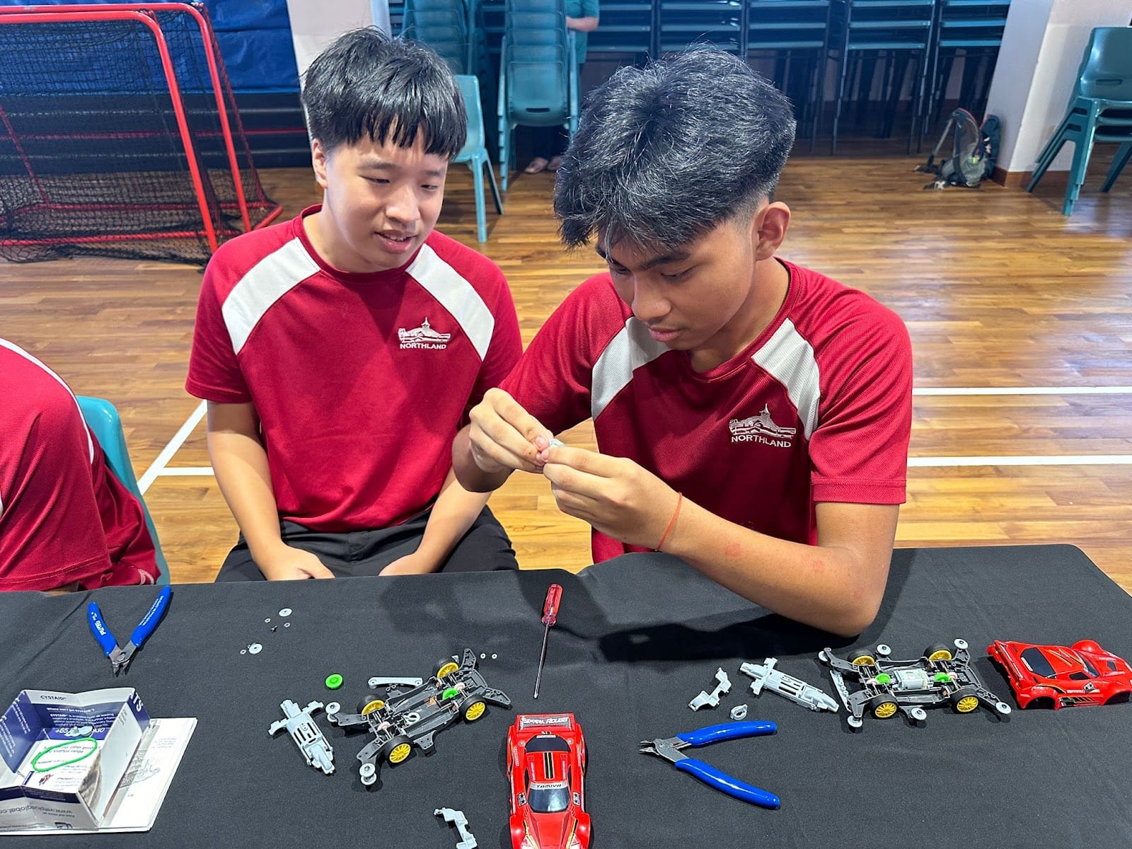 Two boys in red Northland shirts assemble small toy race cars with tools on a black table.