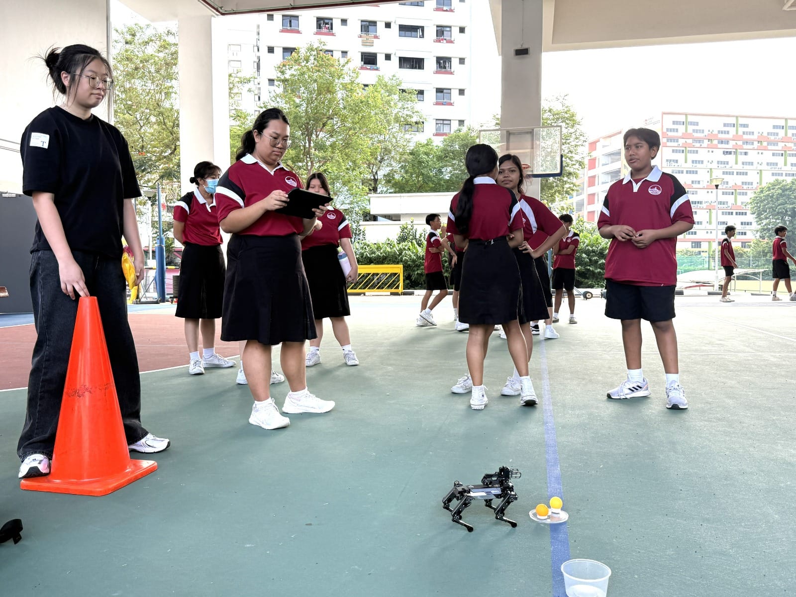 Students in red uniforms stand on a school court near a small robot, orange cone, and balls during an outdoor activity.