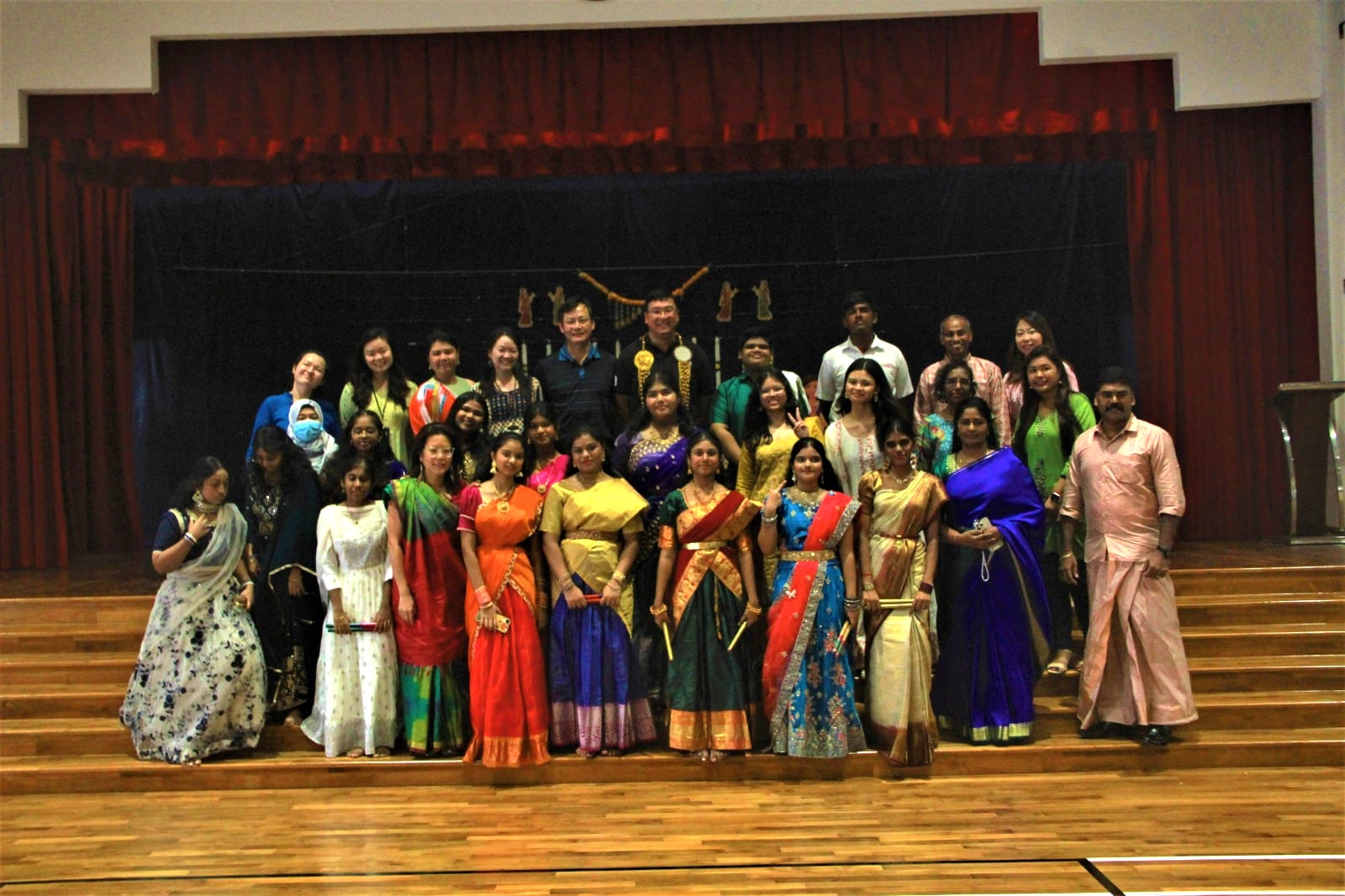 A large group of people posing on a stage, many wearing colorful traditional Indian attire including saris and lehengas.