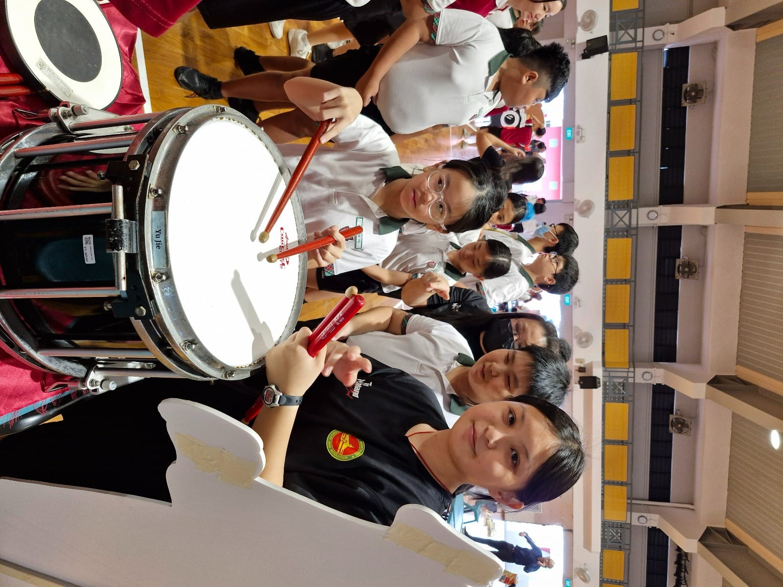 Students in school uniforms hold red drumsticks near a large bass drum indoors.