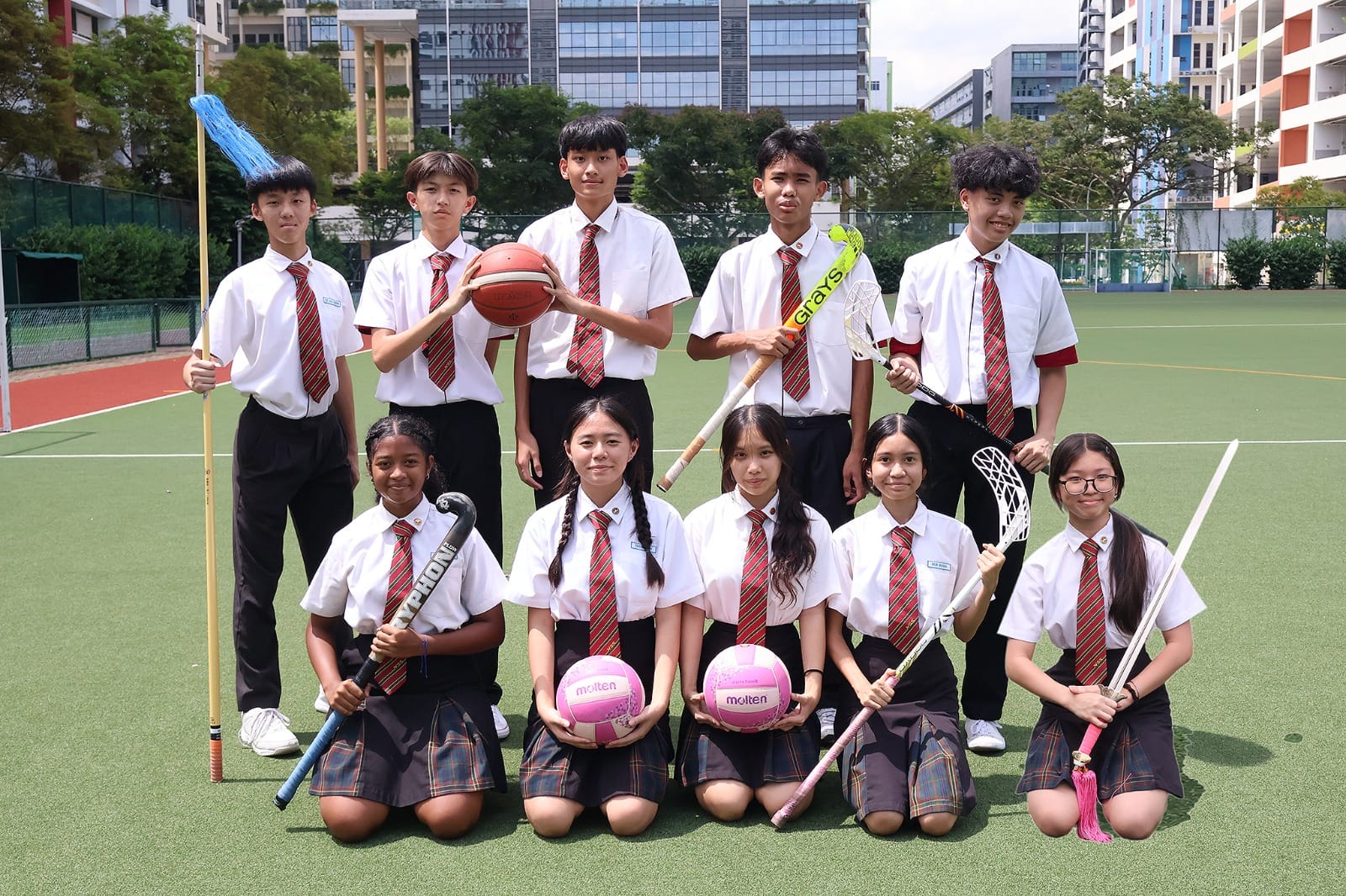 Ten students in white shirts and plaid ties pose on a sports court holding a basketball, hockey sticks, and pink volleyballs.
