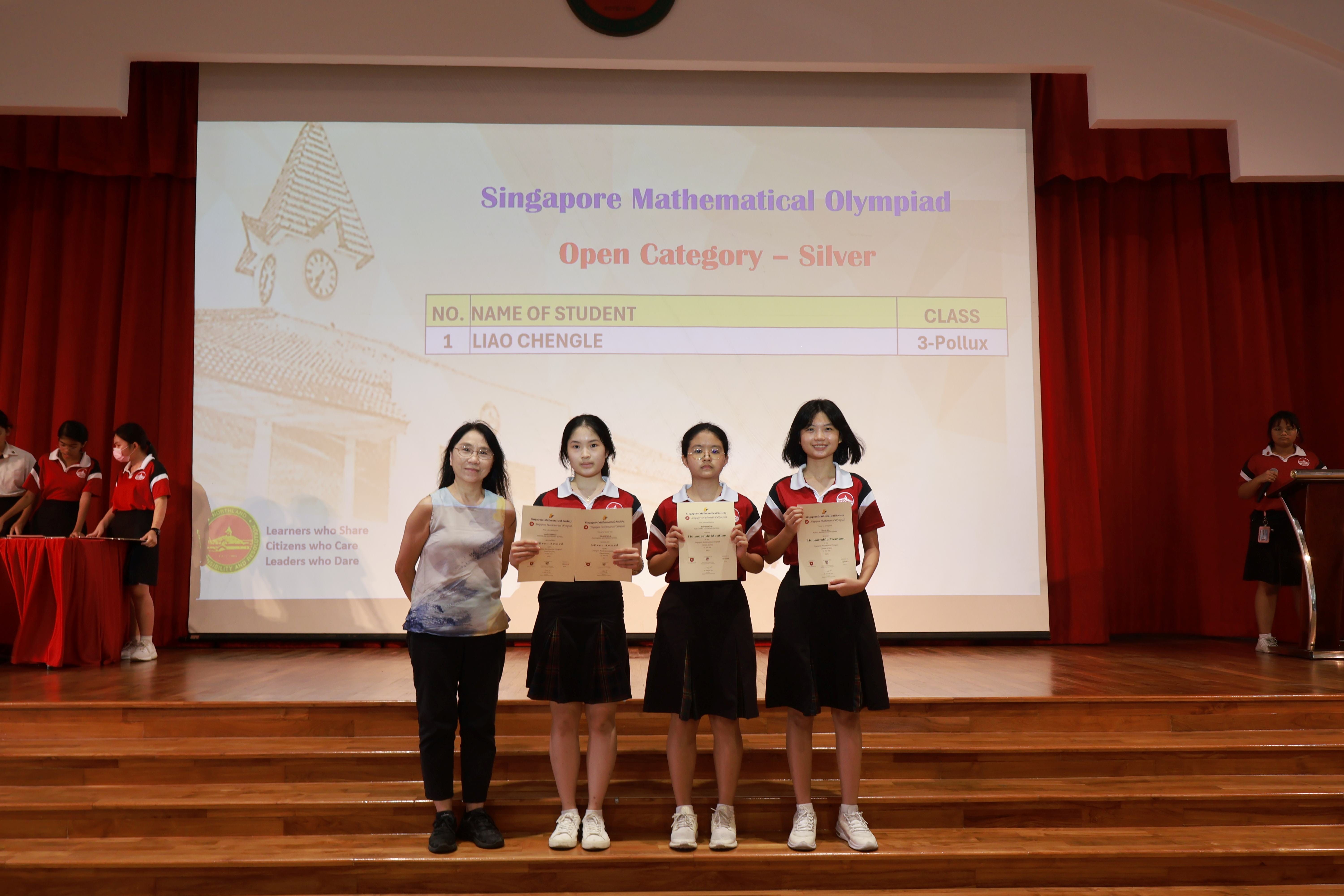 Three students hold certificates on stage with a teacher, in front of a Singapore Mathematical Olympiad Silver award slide.