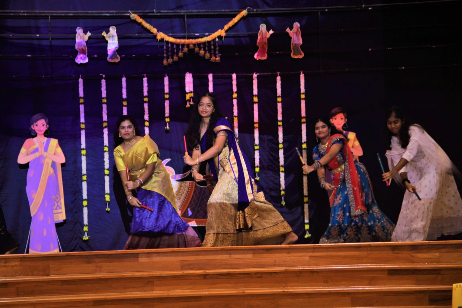 Four women in colorful traditional Indian attire dance with sticks on a decorated stage with floral garlands.