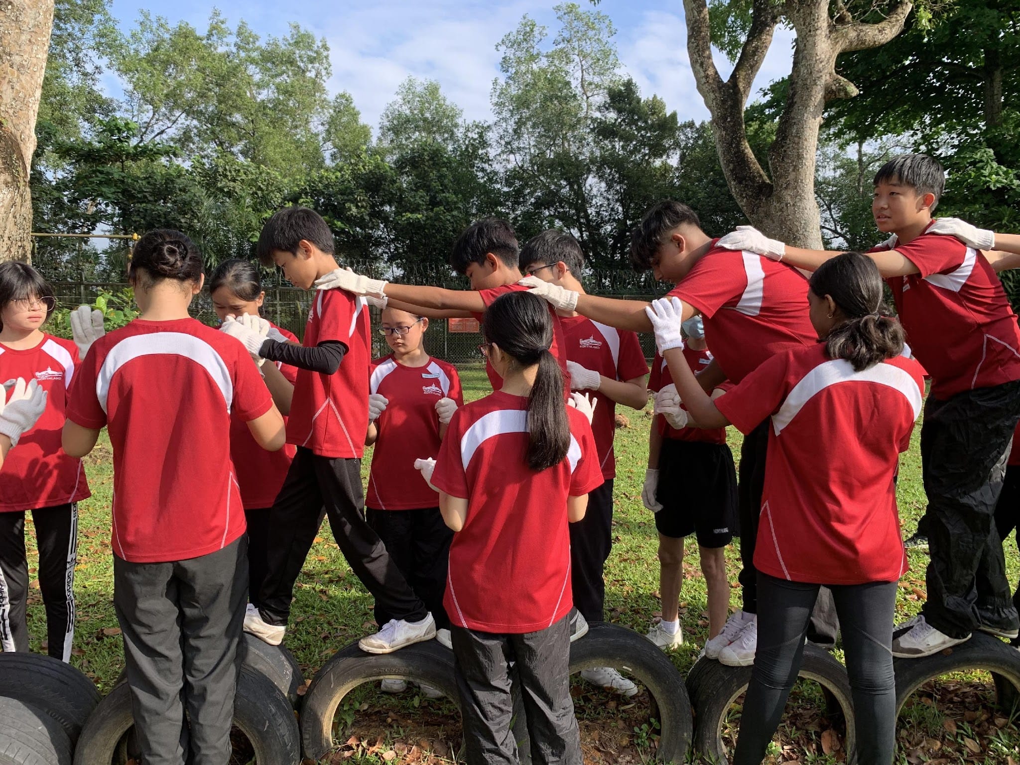 Students in red uniforms with white gloves stand on old tires outdoors, placing hands on each other's shoulders in a circle.