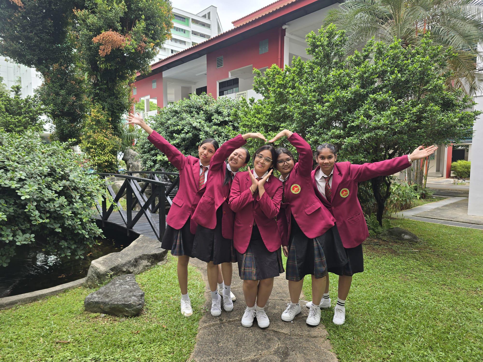 Five girls in matching maroon school blazers pose playfully outdoors near a small bridge and red building.