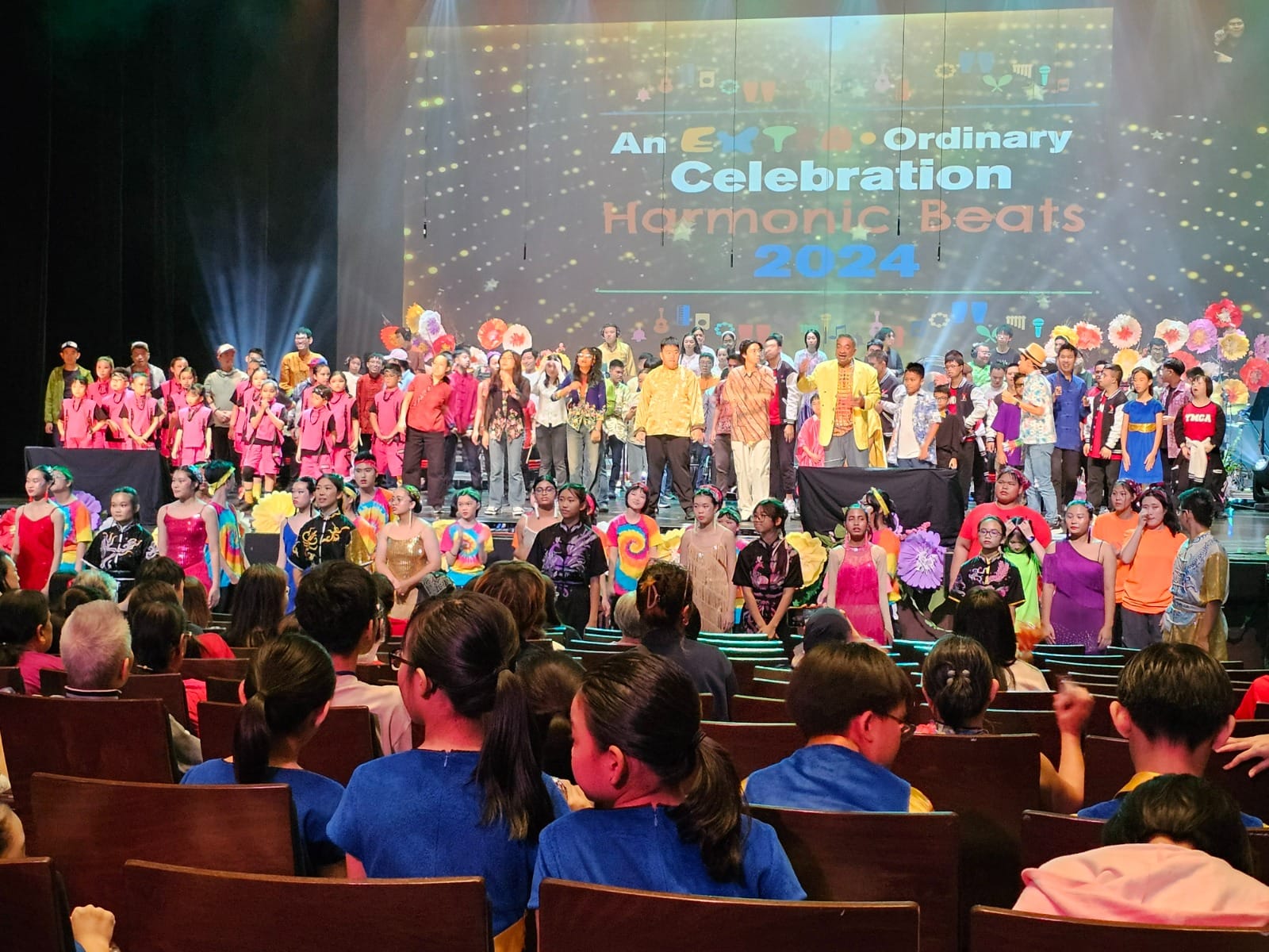 Large group of children and adults in colorful costumes on stage at Harmonic Beats 2024 celebration event.