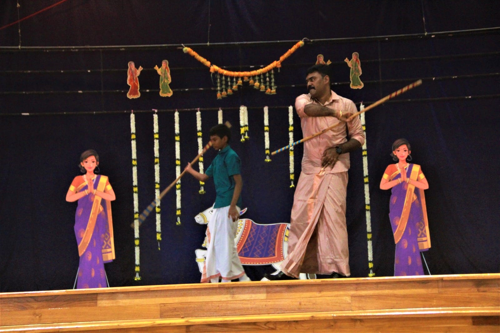 Two people holding sticks perform on a decorated stage with cardboard cutouts of women and a bull.