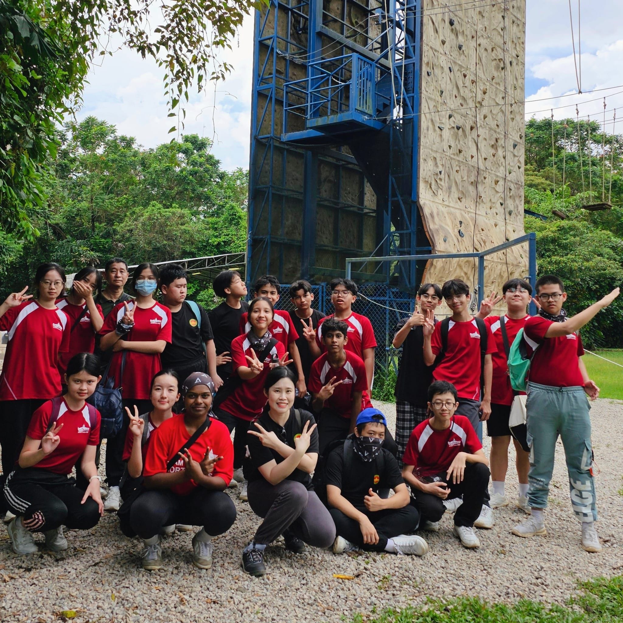 A group of young people in red shirts pose with peace signs in front of a tall outdoor rock climbing wall.