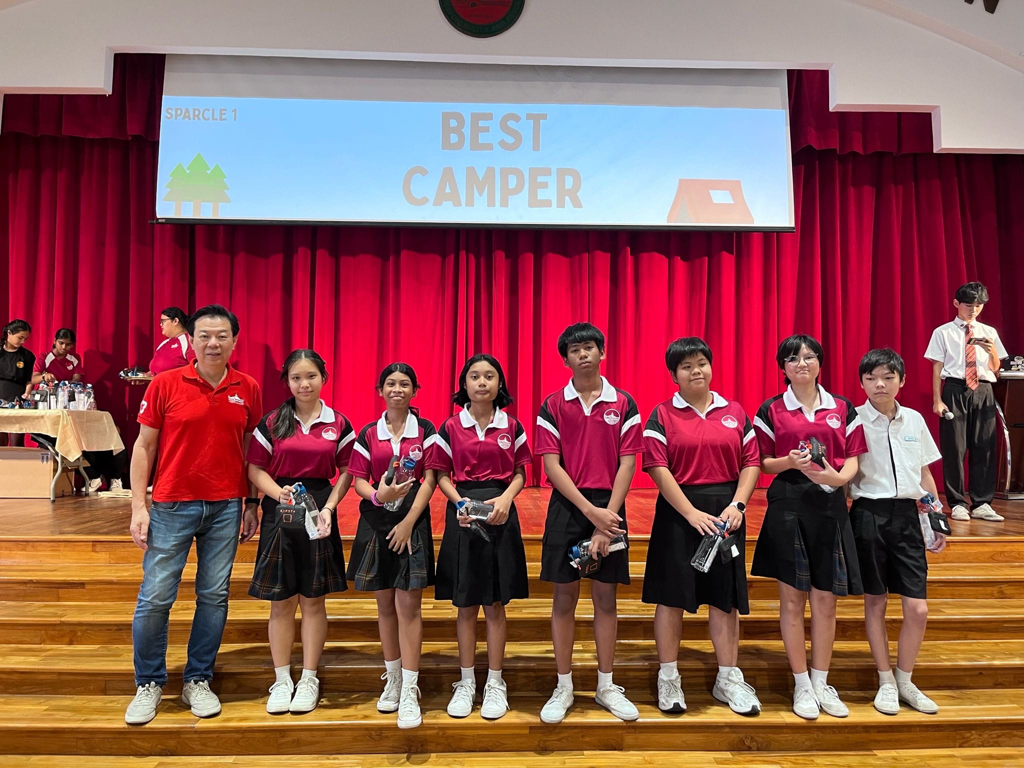 A man and six students in red uniforms stand on a stage in front of a Best Camper screen with red curtains.