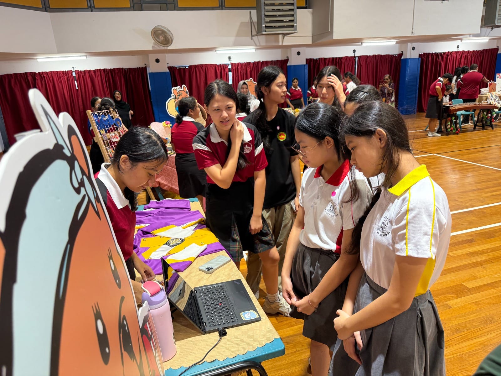 Students in school uniforms gather around a booth with a laptop in a school hall during a fair.