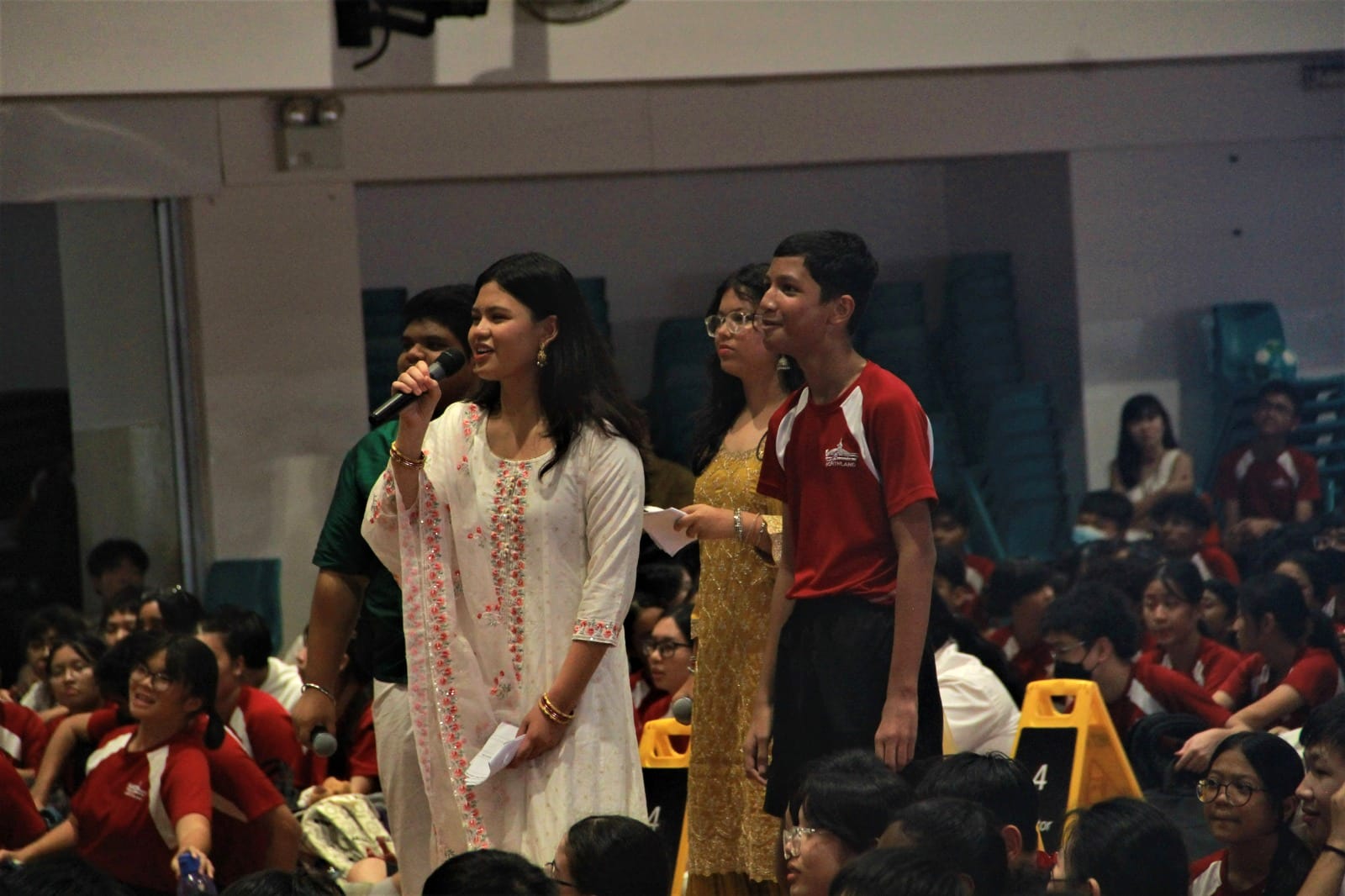 A girl in a white embroidered outfit speaks into a microphone at a school event, surrounded by students in red uniforms.