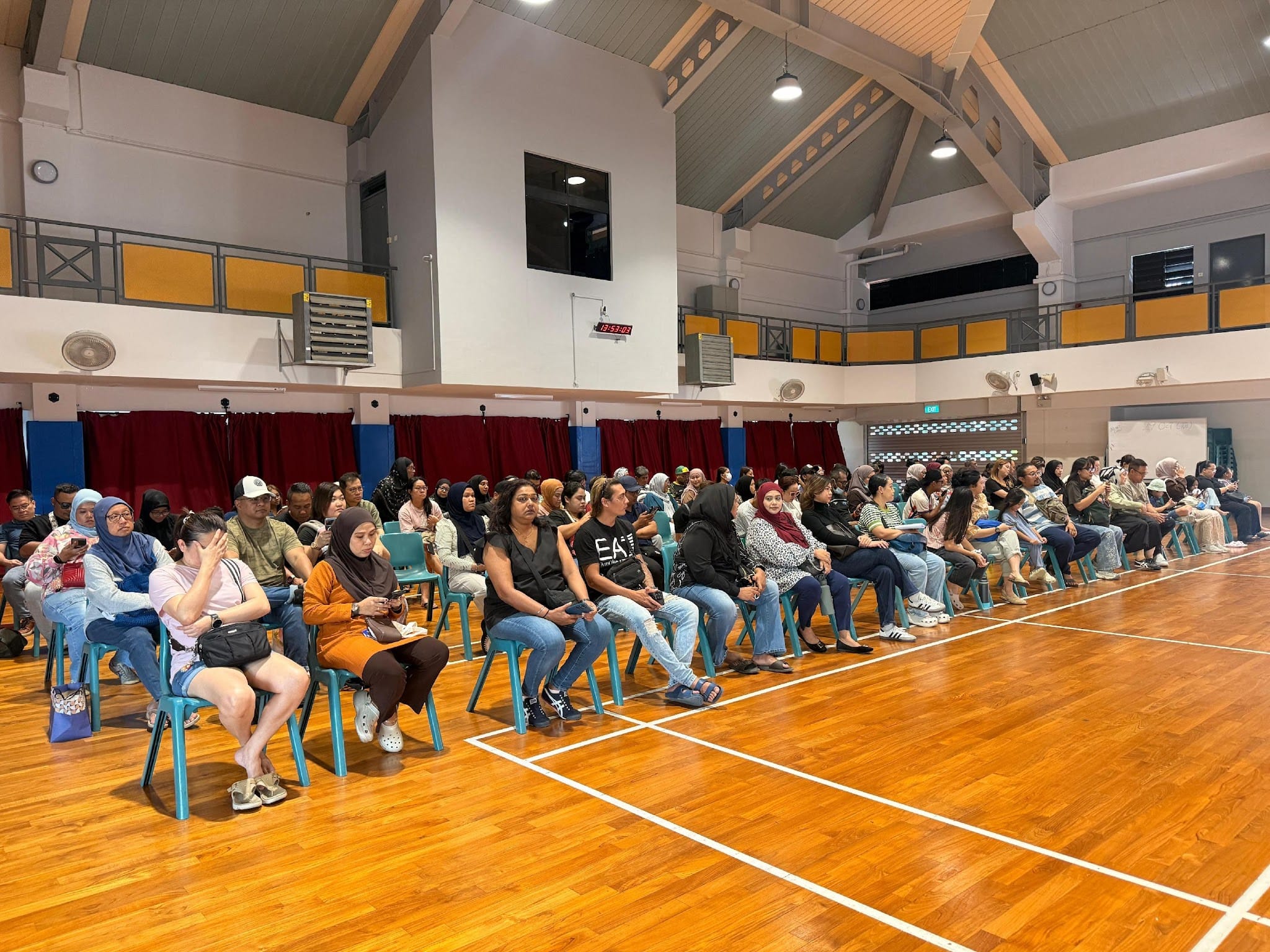 A large group of adults seated on teal chairs in rows inside a school gymnasium with a wooden floor.