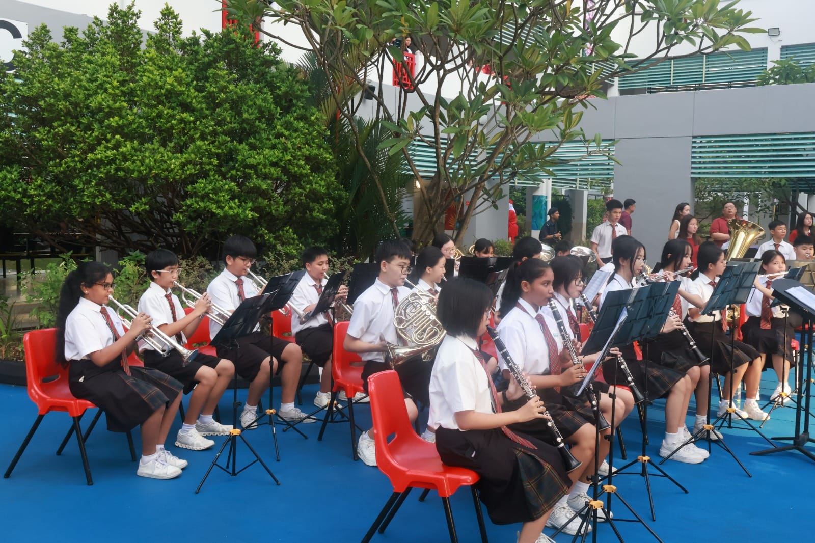 Students in school uniforms sit on red chairs outdoors playing brass and woodwind instruments with sheet music stands.